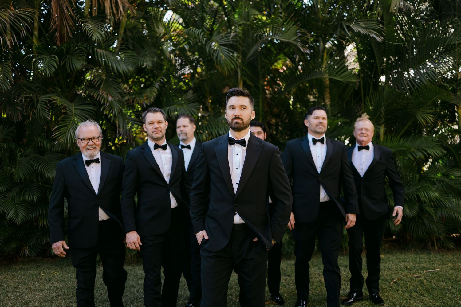 Groom and groomsmen in matching black tuxedos posing confidently in front of lush tropical palm foliage.
