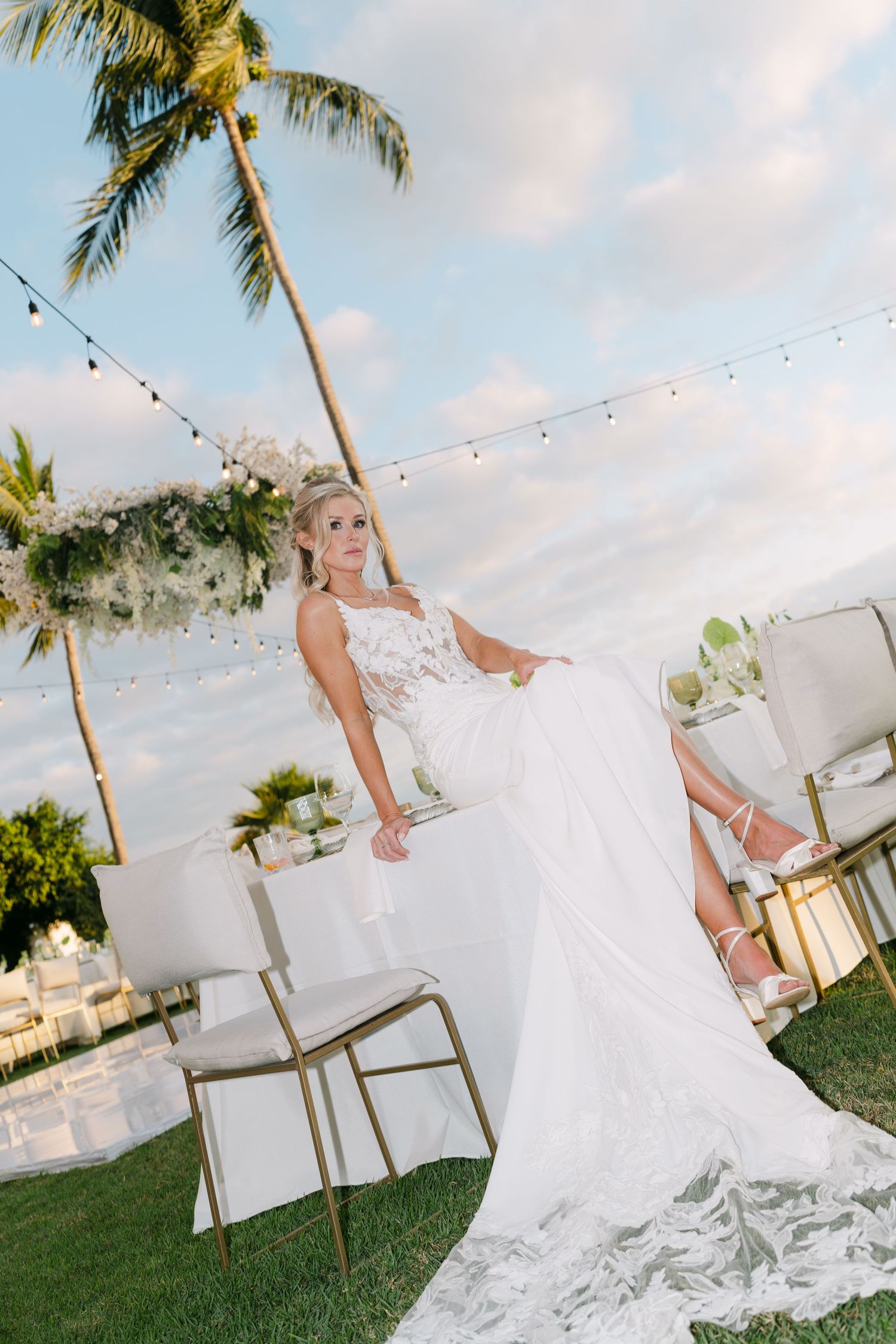 Bride in elegant white lace gown posing beside sweetheart table under palm tree with string lights at sunset.