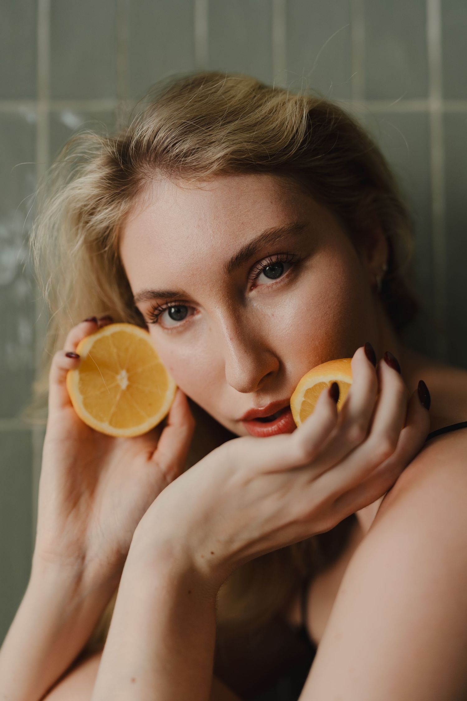 Blonde woman holding two halved oranges against her face in moody green-tiled bathroom portrait.