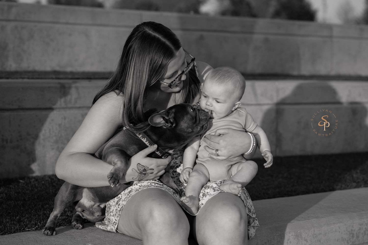 Black and white photo of a woman holding a baby and a small dog, sharing a tender moment outdoors.