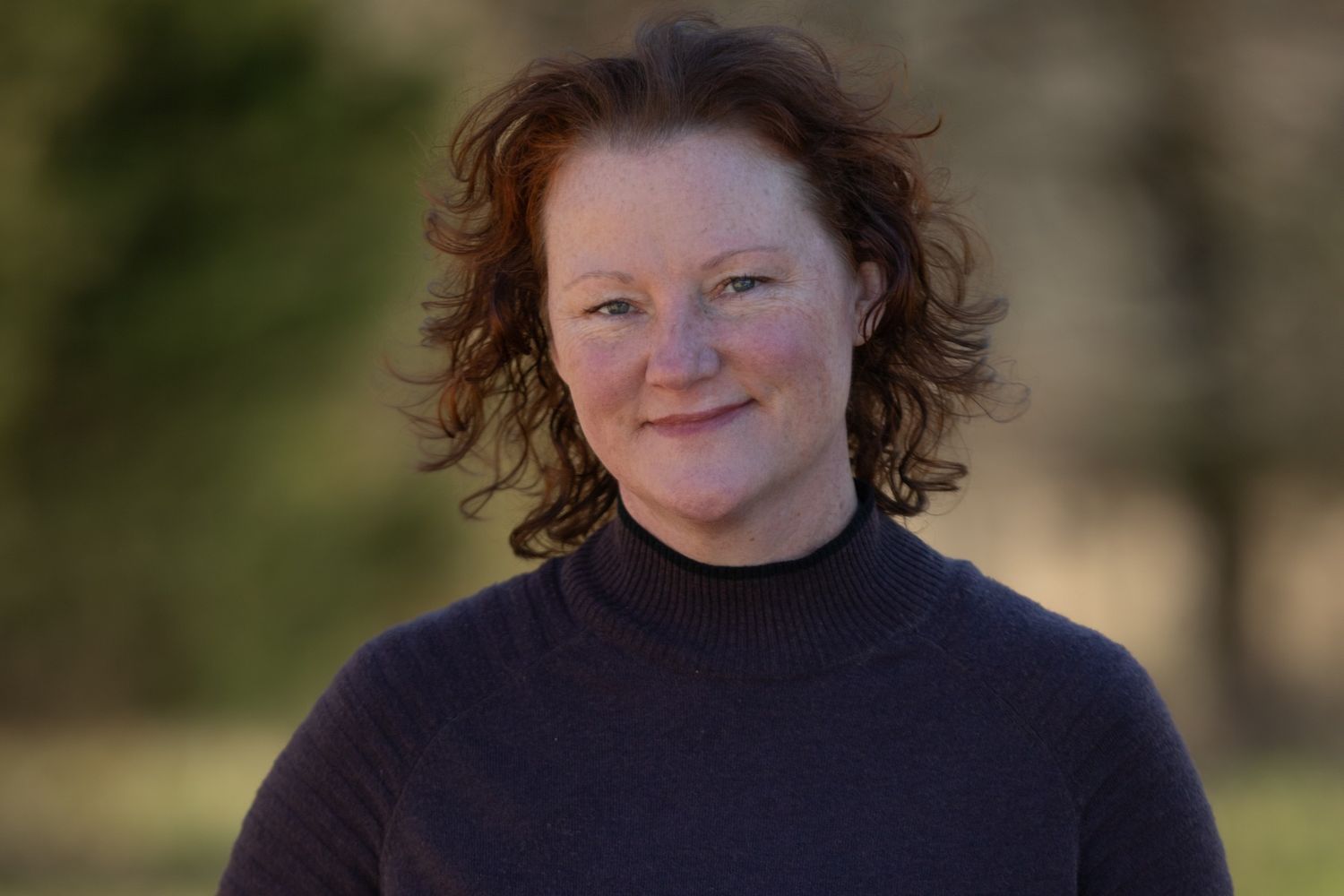 A smiling woman with curly red hair wearing a dark turtleneck sweater outdoors with a blurred green background.