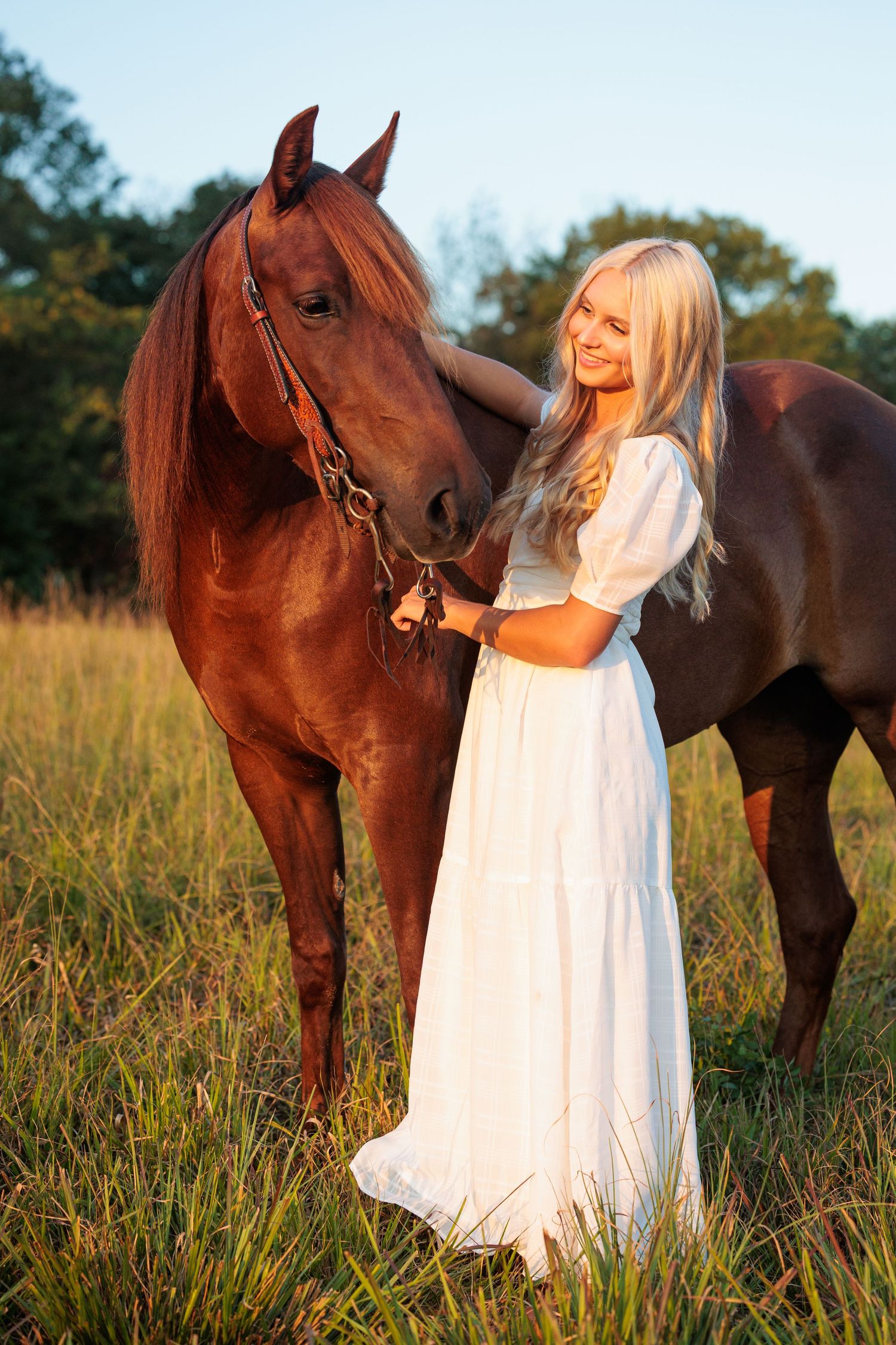 A person in a flowing white dress stands in a grassy field with a beautiful chestnut horse at sunset.