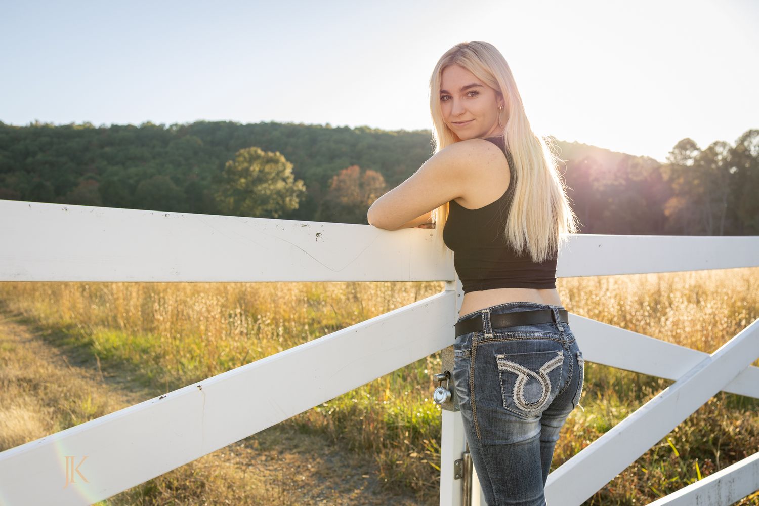 Blonde woman in black tank top and jeans leaning on white fence at golden hour on a sunny farm.