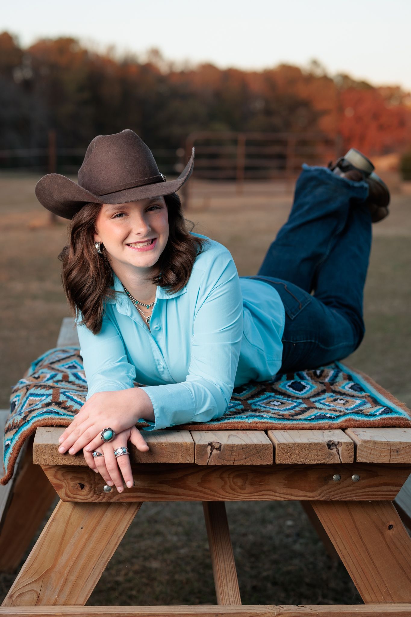 Young woman in cowboy hat and turquoise shirt poses on wooden bench with colorful blanket in rural outdoor setting.