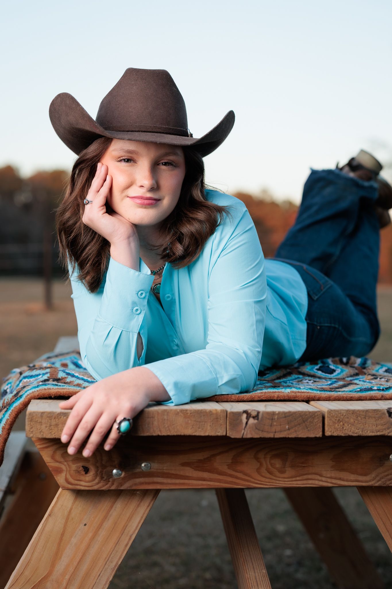 Young girl in turquoise shirt and brown cowboy hat poses on wooden bench outdoors.
