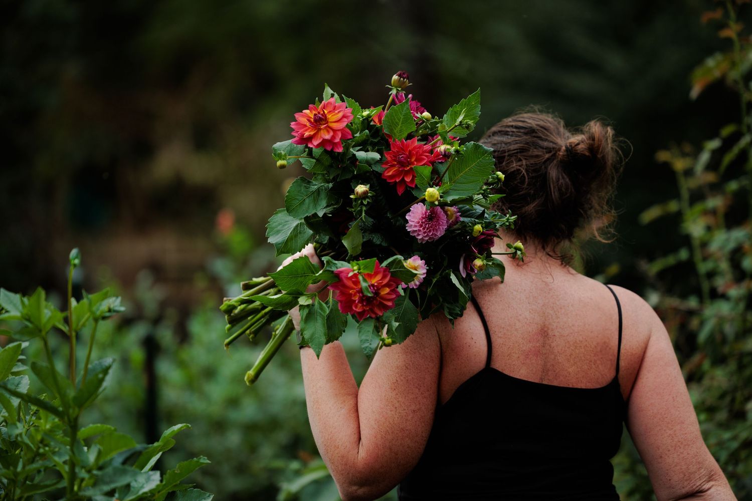 Person in black top carrying vibrant red and pink floral arrangement through a lush garden.