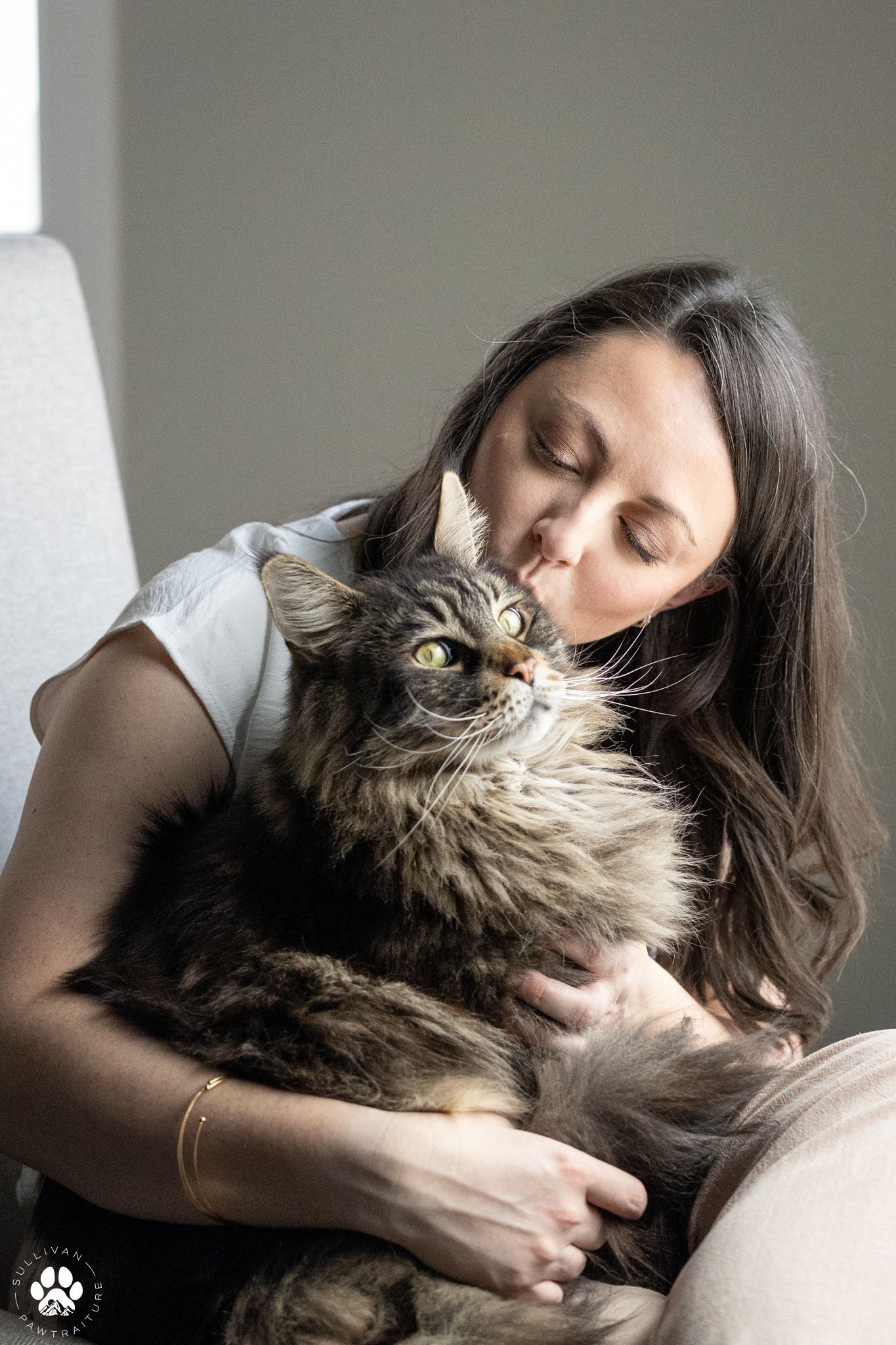 A woman with long dark hair cuddles a fluffy Maine Coon cat while seated in a chair.