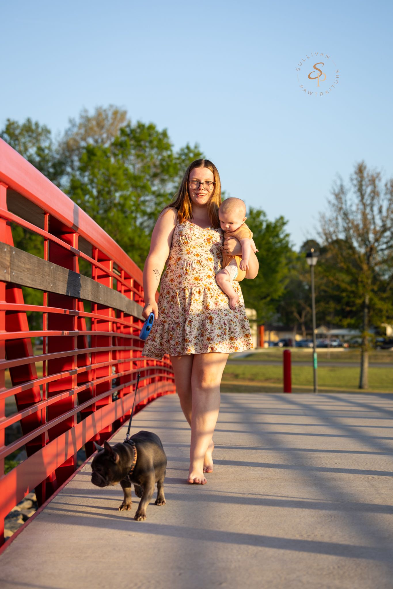 Woman in floral dress holds baby while walking miniature pig on red bridge in sunny park.