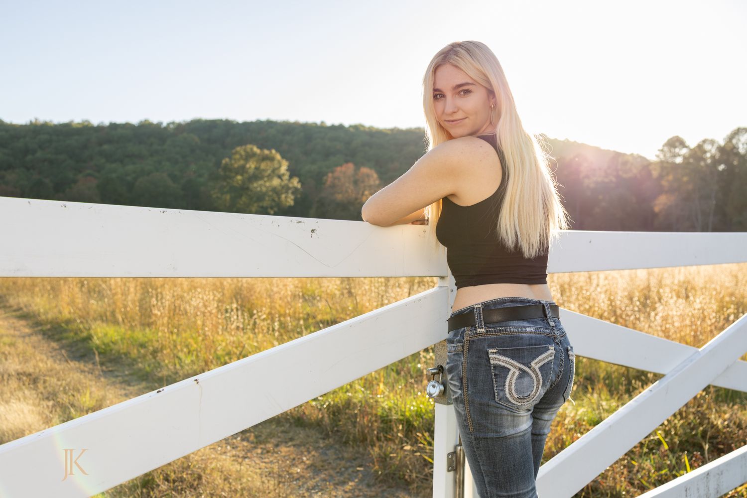 Young blonde woman in black tank top and jeans leaning on white fence at golden hour on a sunny farm.
