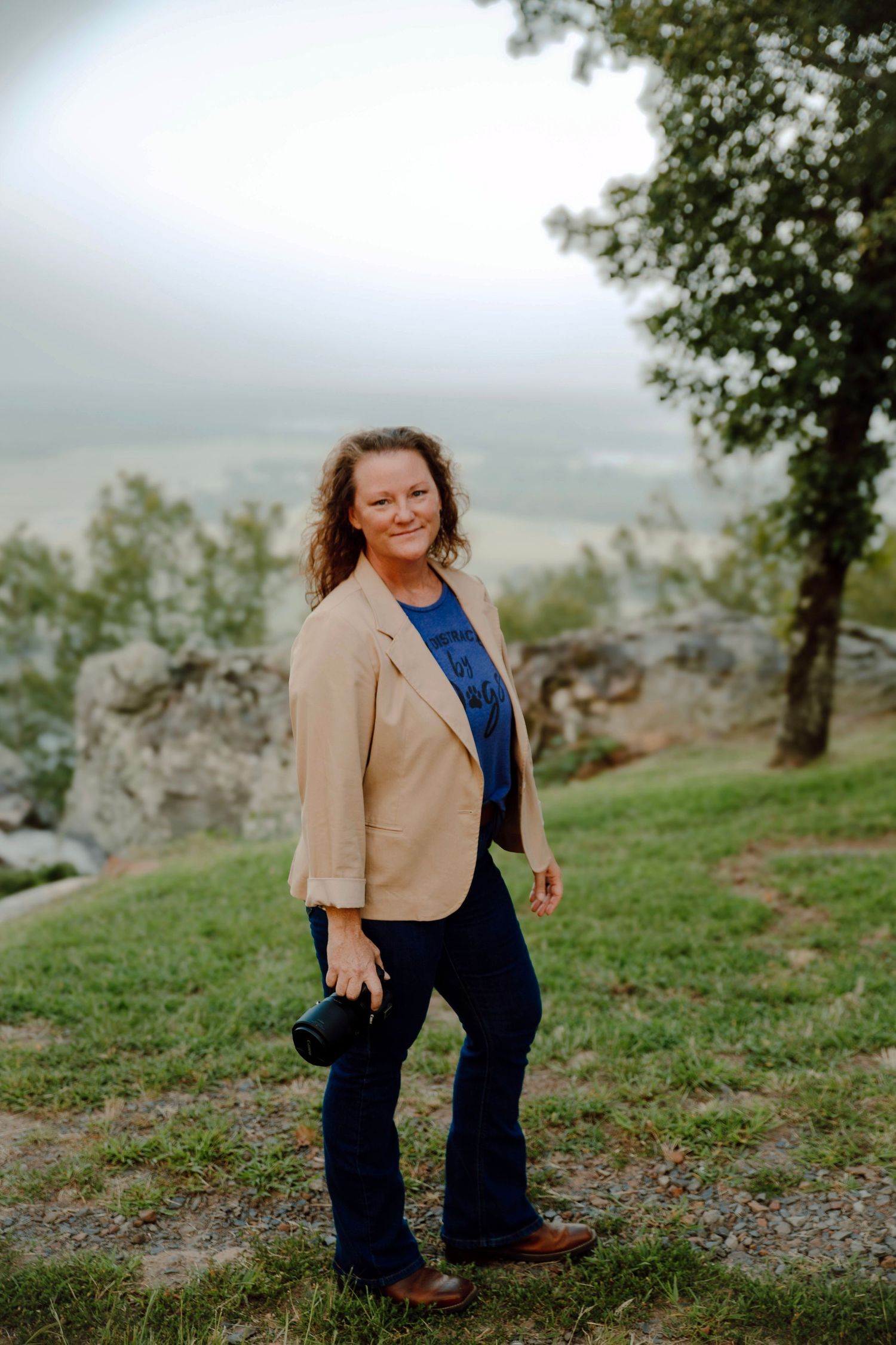 A person in a tan blazer and blue top poses on a scenic overlook with rocky terrain and trees in the background.