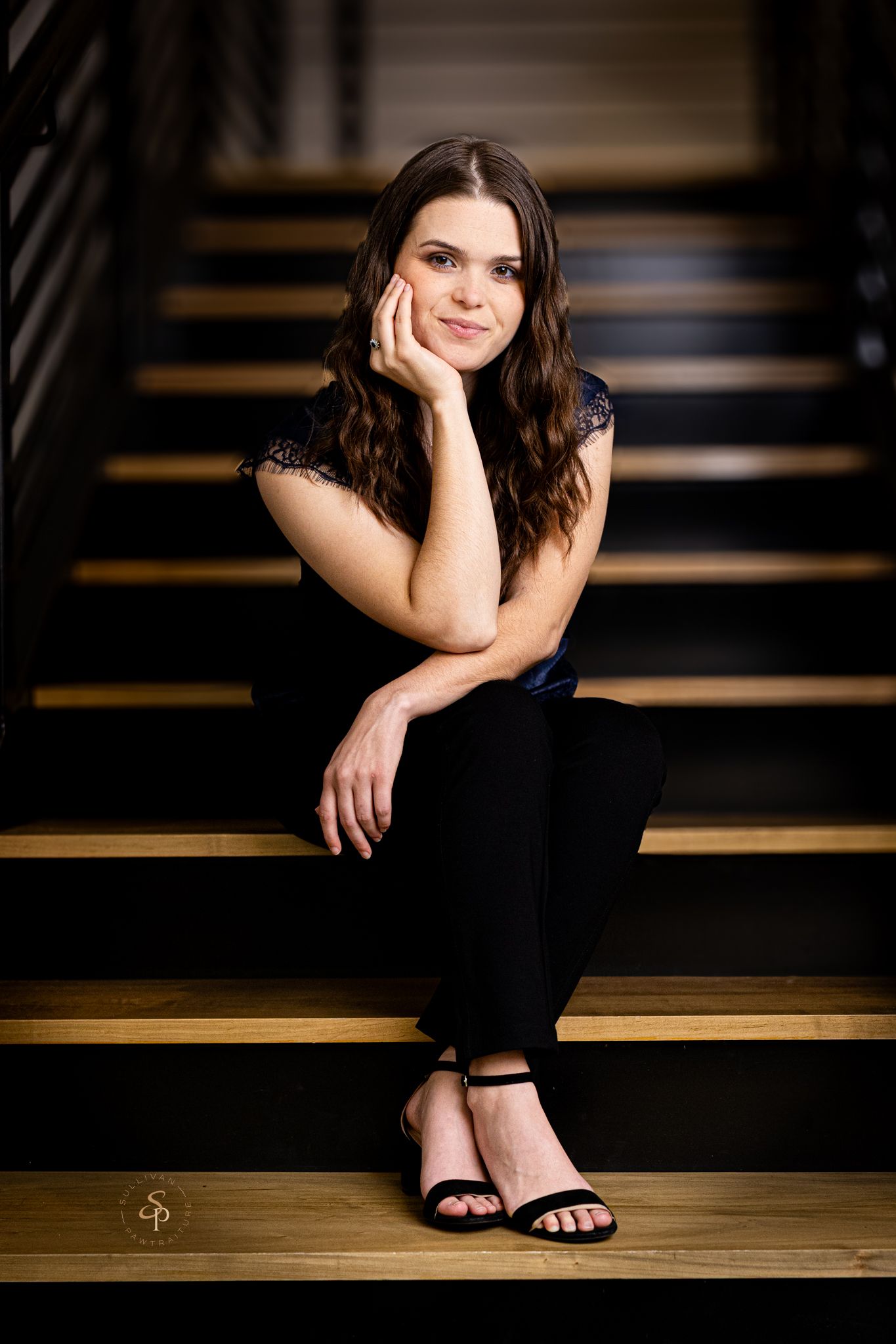 Young woman in black outfit sits gracefully on wooden stairs, resting chin on hand, smiling softly at camera.