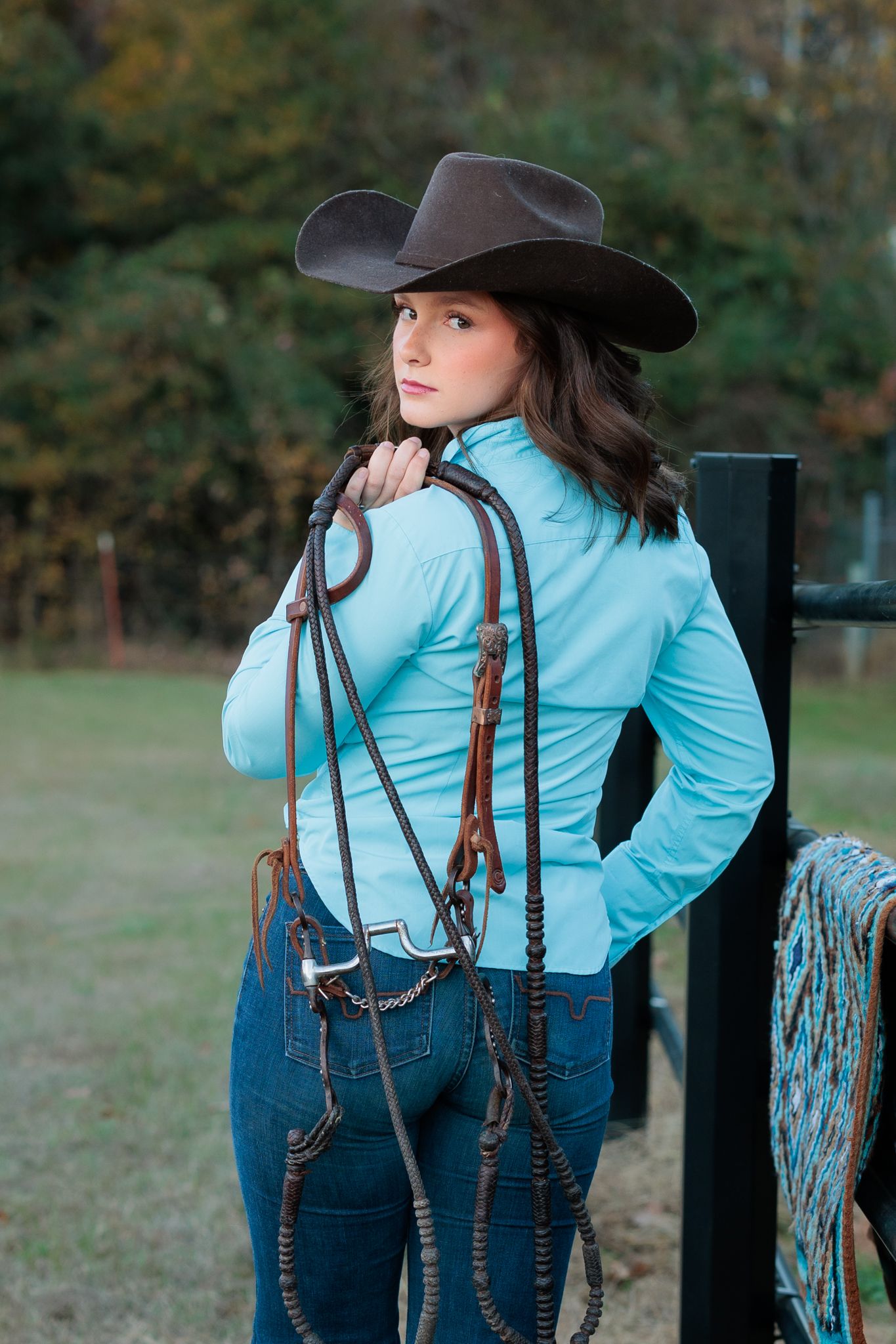 Young cowgirl in teal jacket and brown hat holds horse reins while leaning on a fence outdoors.