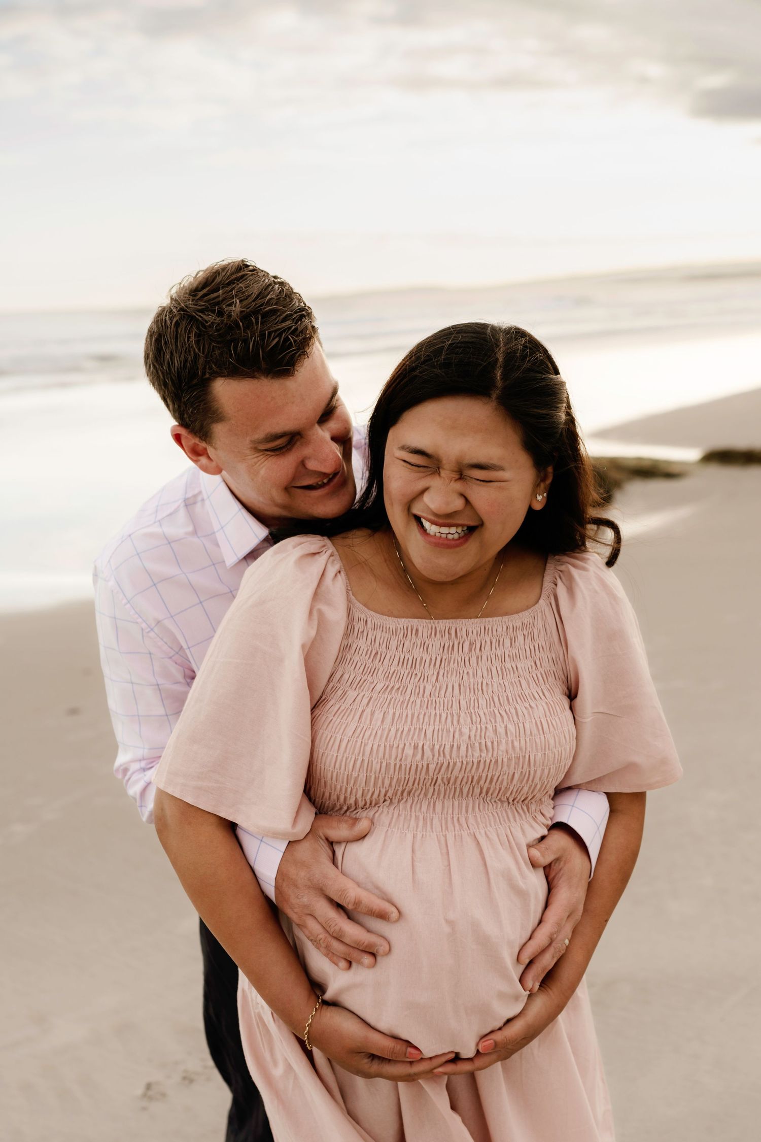 Couple sharing a joyful moment on the beach, with pregnant woman in pink dress laughing as partner embraces her from behind.
