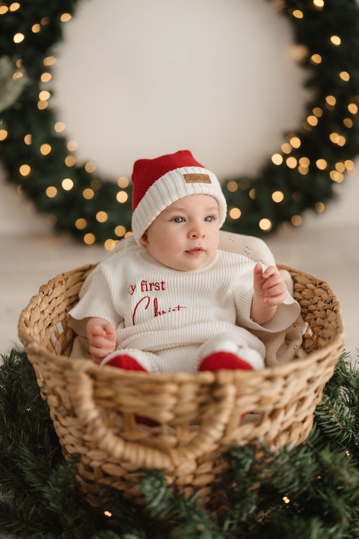 Baby wearing white sweater and winter hat lies in woven basket with Christmas decorations and twinkling lights in background.