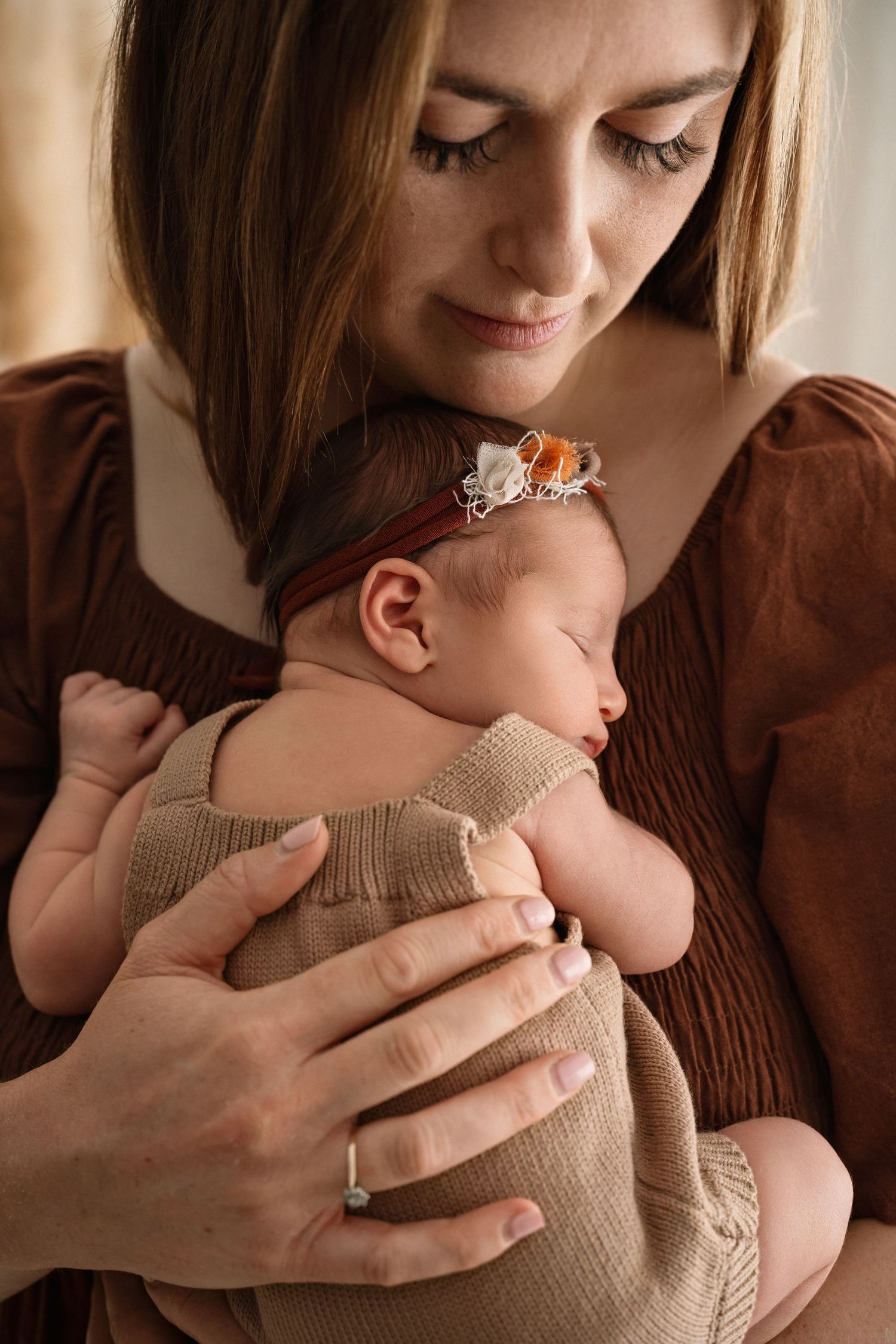 A mother in a brown dress tenderly holds her sleeping newborn baby wrapped in a beige swaddle with a floral headband.