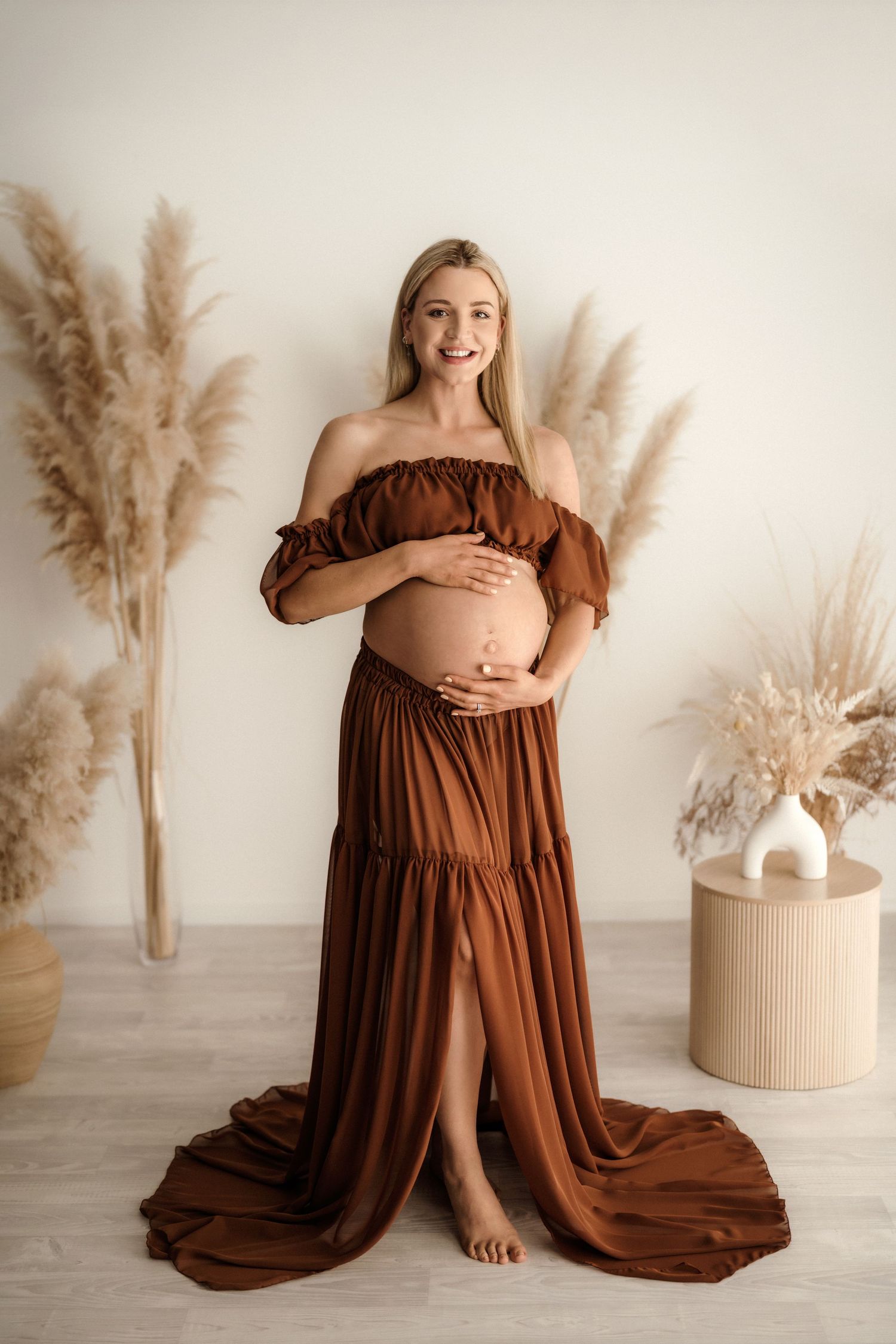 Smiling pregnant woman in rust off-shoulder maternity gown holding belly, posed with pampas grass decor.