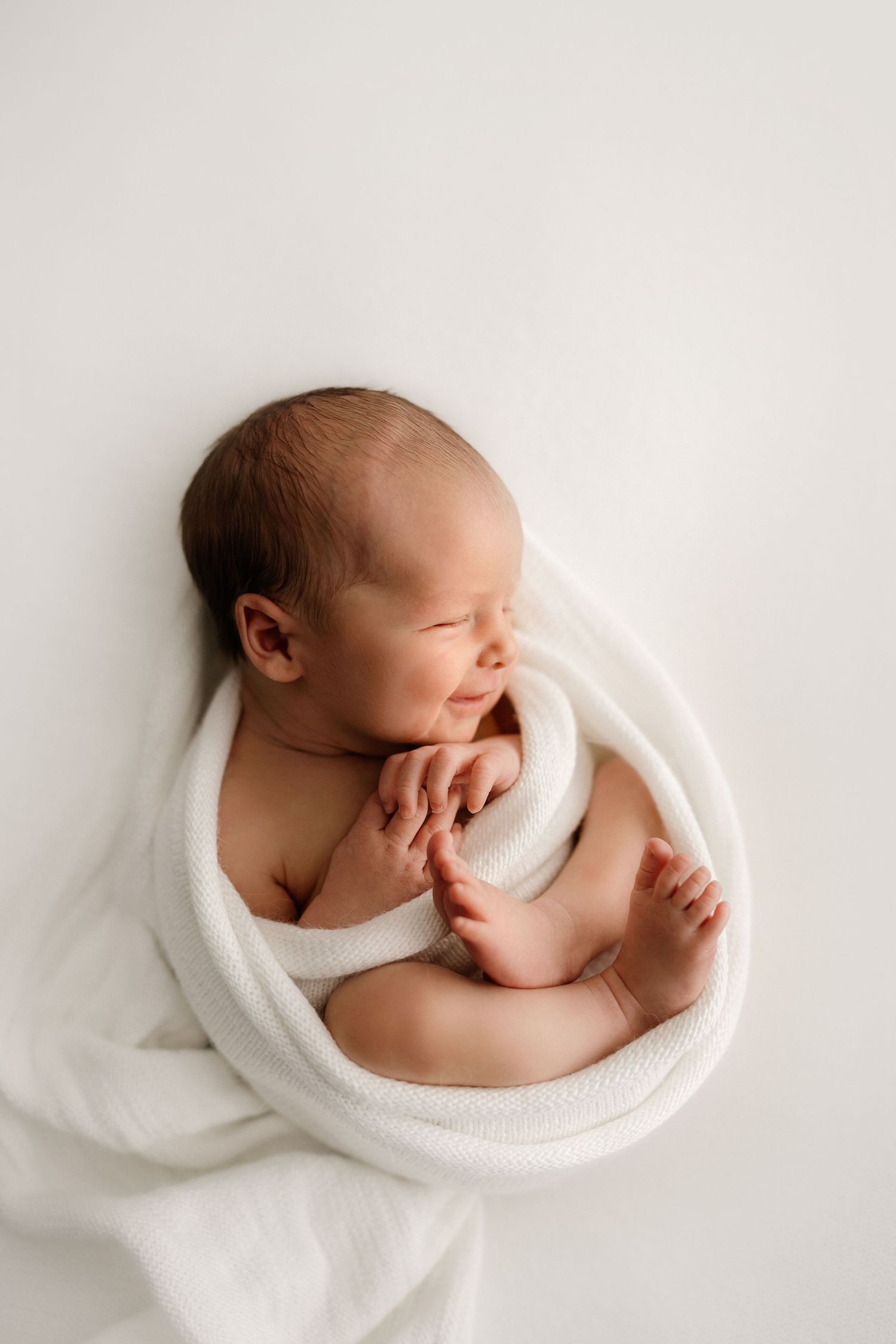 Peaceful newborn baby swaddled in soft white blanket sleeps with tiny hands curled during professional photo session.