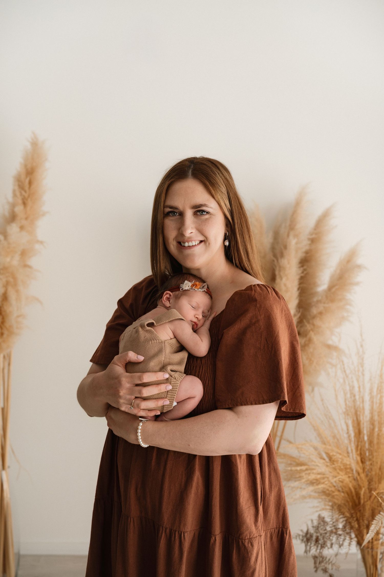 Smiling woman in rust dress holding sleeping newborn baby, surrounded by pampas grass in a soft studio setting.