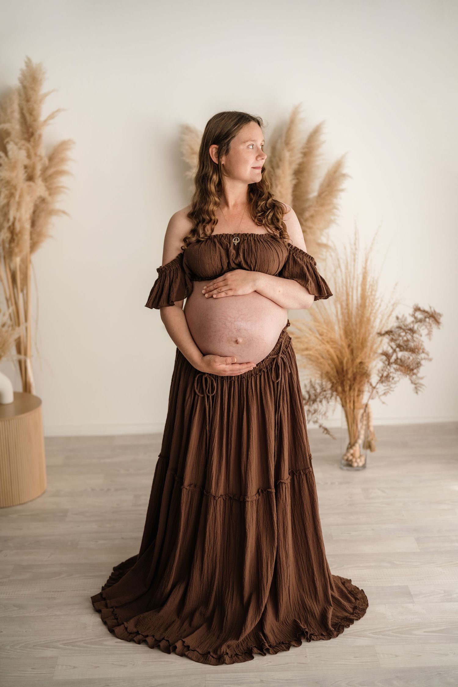 Pregnant woman in brown off-shoulder dress cradling baby bump, surrounded by pampas grass in a boho maternity photoshoot.