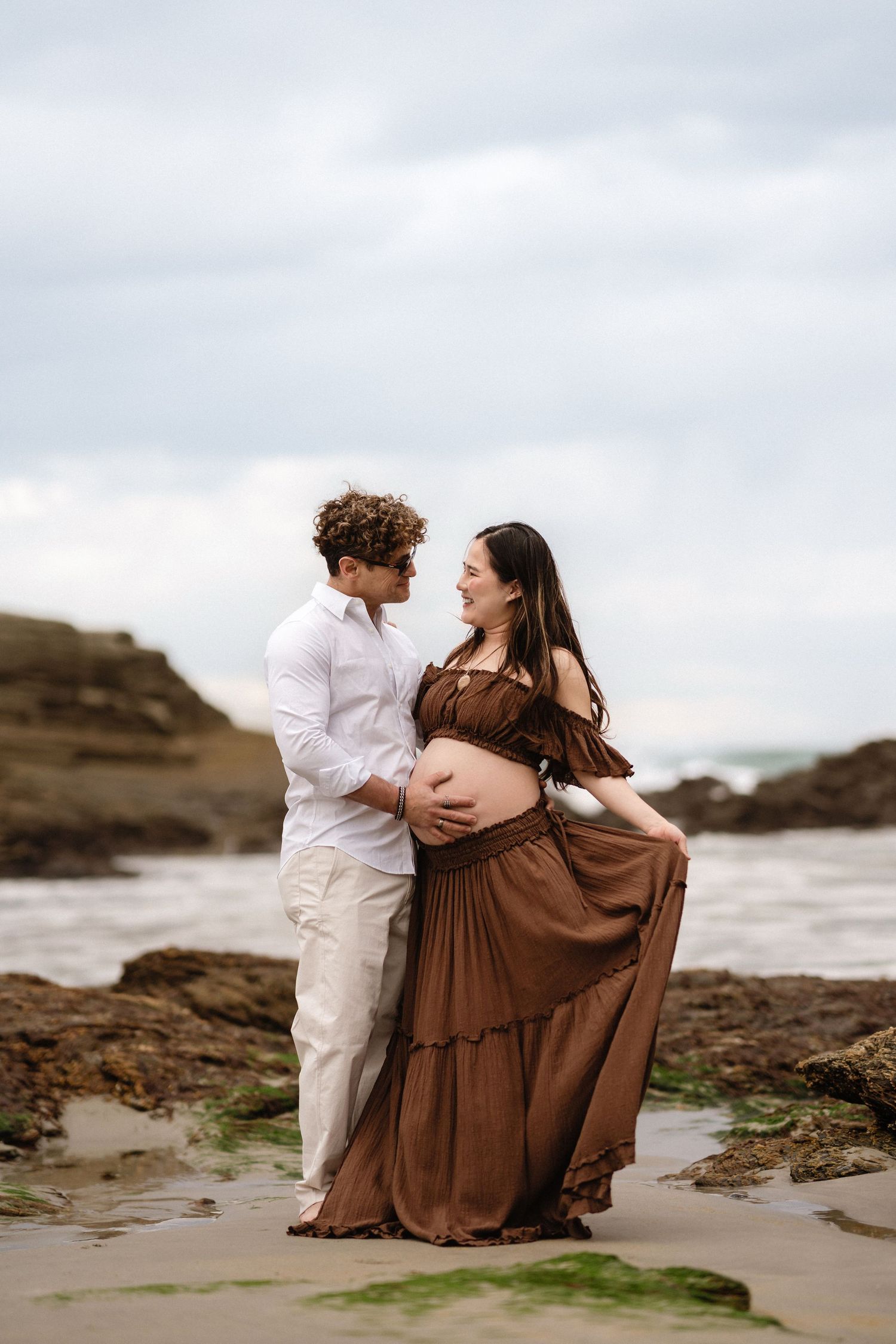 A couple shares a tender moment on a rocky beach, the pregnant woman in a flowing brown dress cradling her belly.