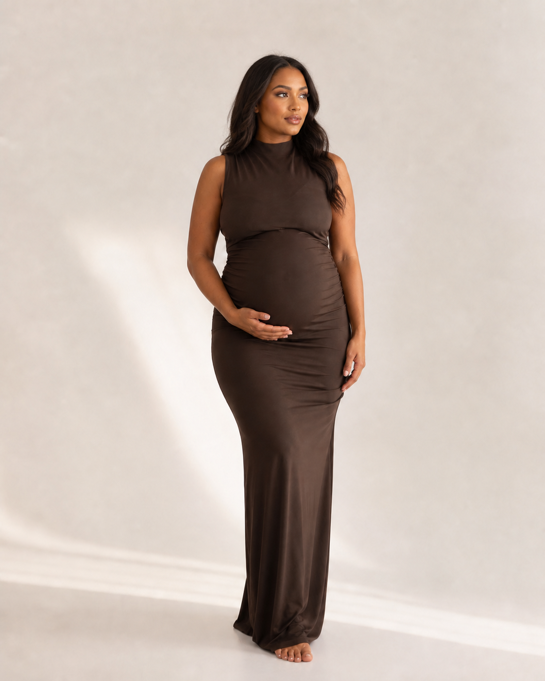 Pregnant woman wearing a sleeveless brown mock neck maxi dress, posing gracefully while cradling her baby bump.