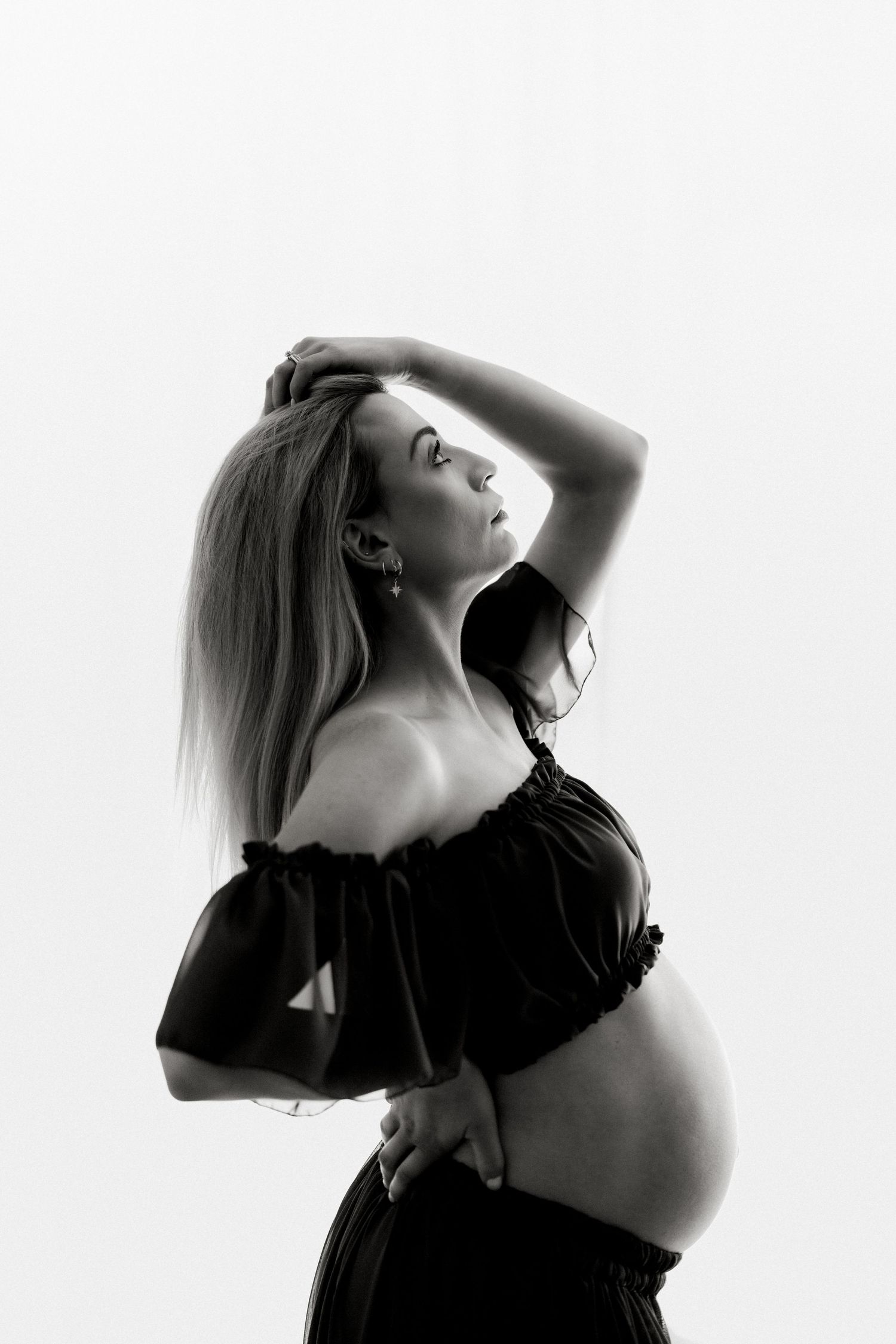 Black and white maternity photo of pregnant woman in off-shoulder top posing gracefully against white background.