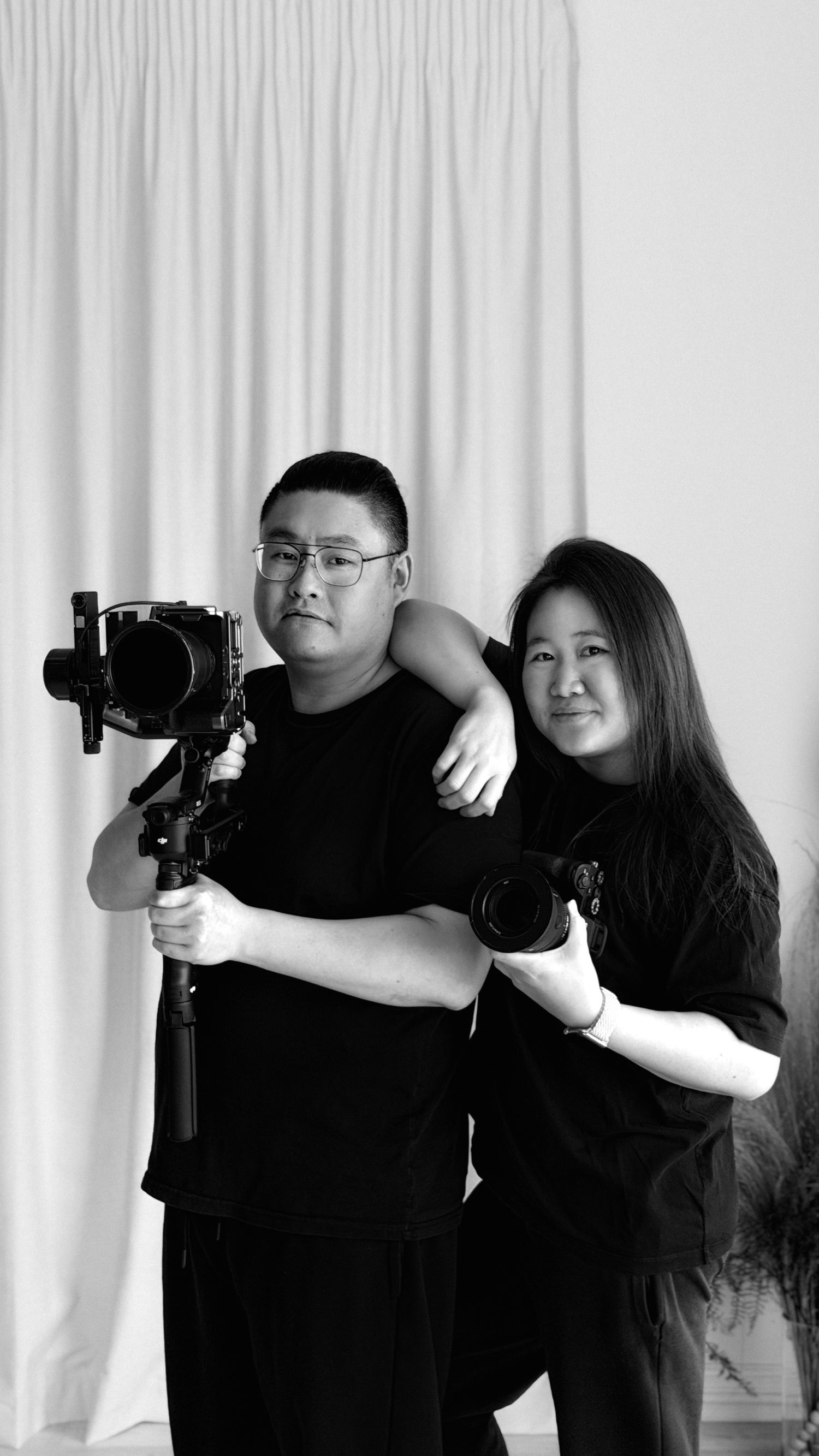 Black and white photo of two photographers posing with a camera gimbal and DSLR camera against a white curtain backdrop.