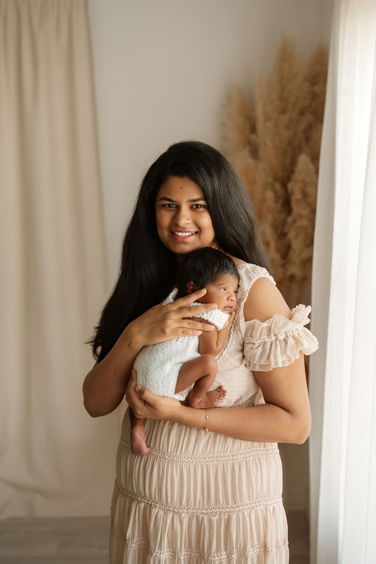 Smiling woman in beige dress holding a sleeping newborn baby, standing in a softly lit studio with pampas grass decor.