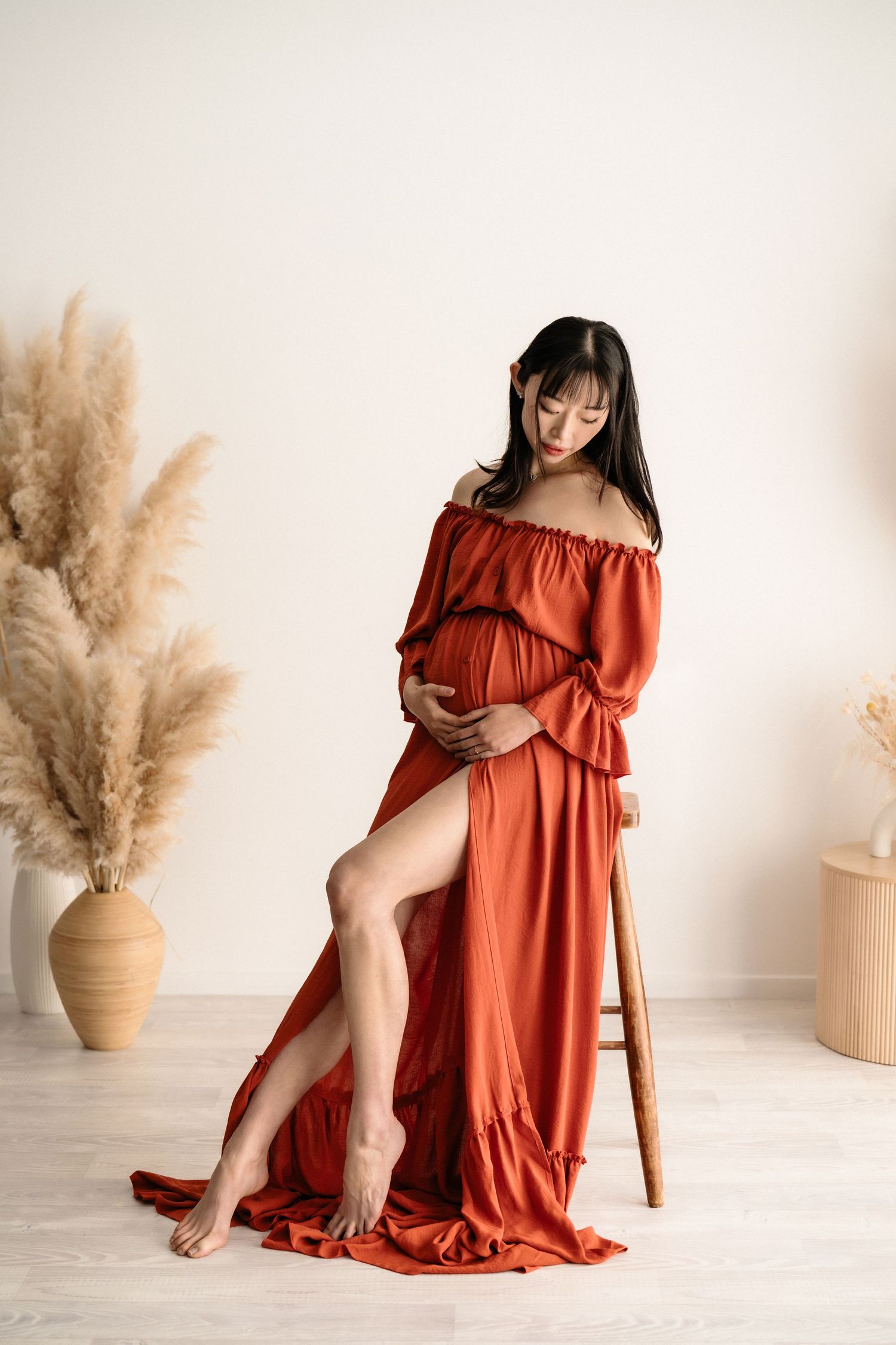 A maternity photo featuring a flowing rust-colored off-shoulder dress against a neutral backdrop with pampas grass decor.