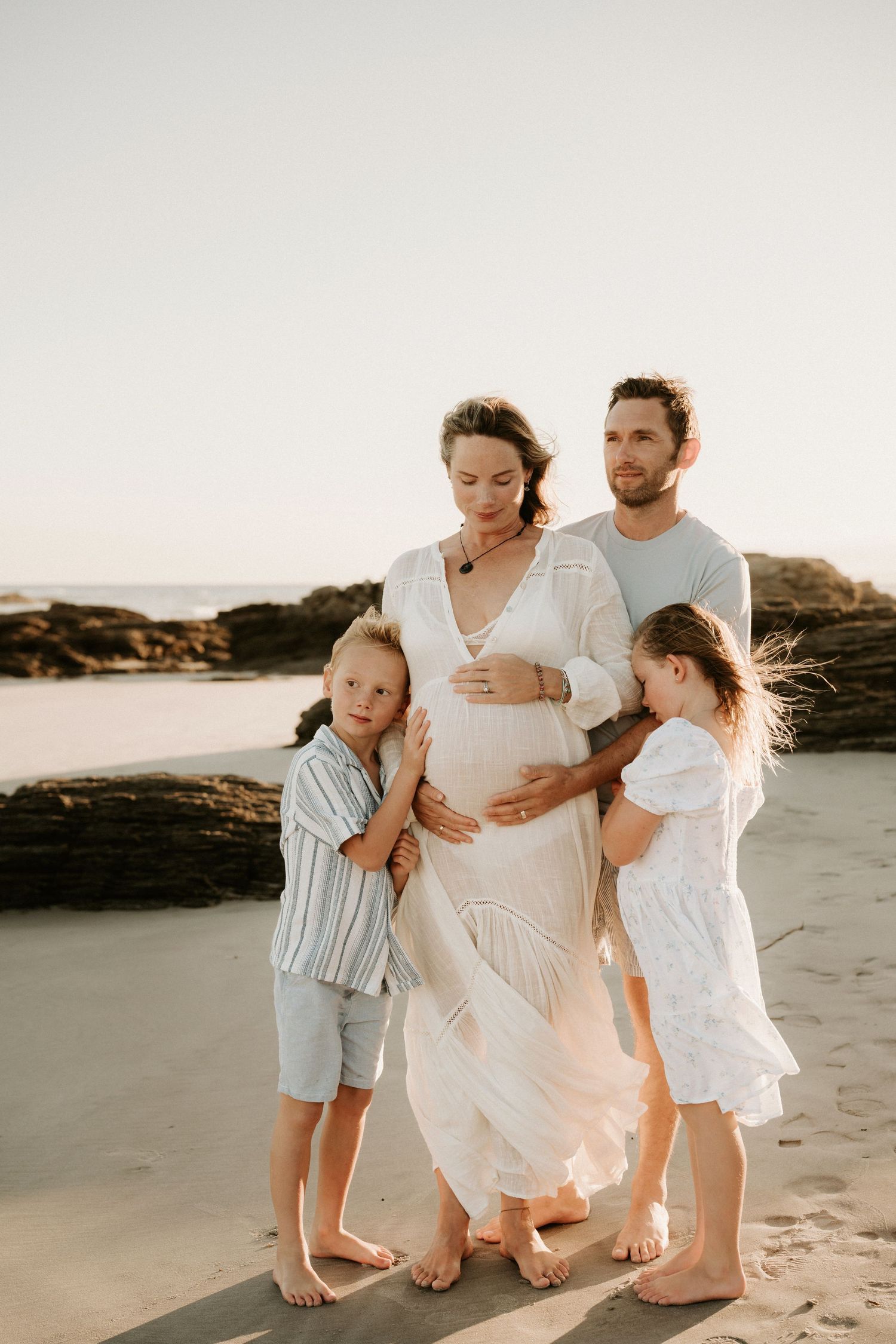Pregnant woman in white dress with husband and two children embracing her baby bump on a sunny beach.