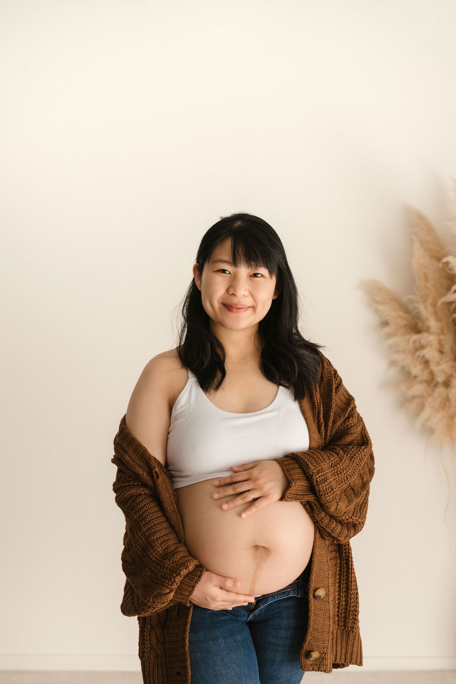 An expectant mother in a white tank top and brown cardigan cradles her baby bump against a neutral wall with pampas grass.