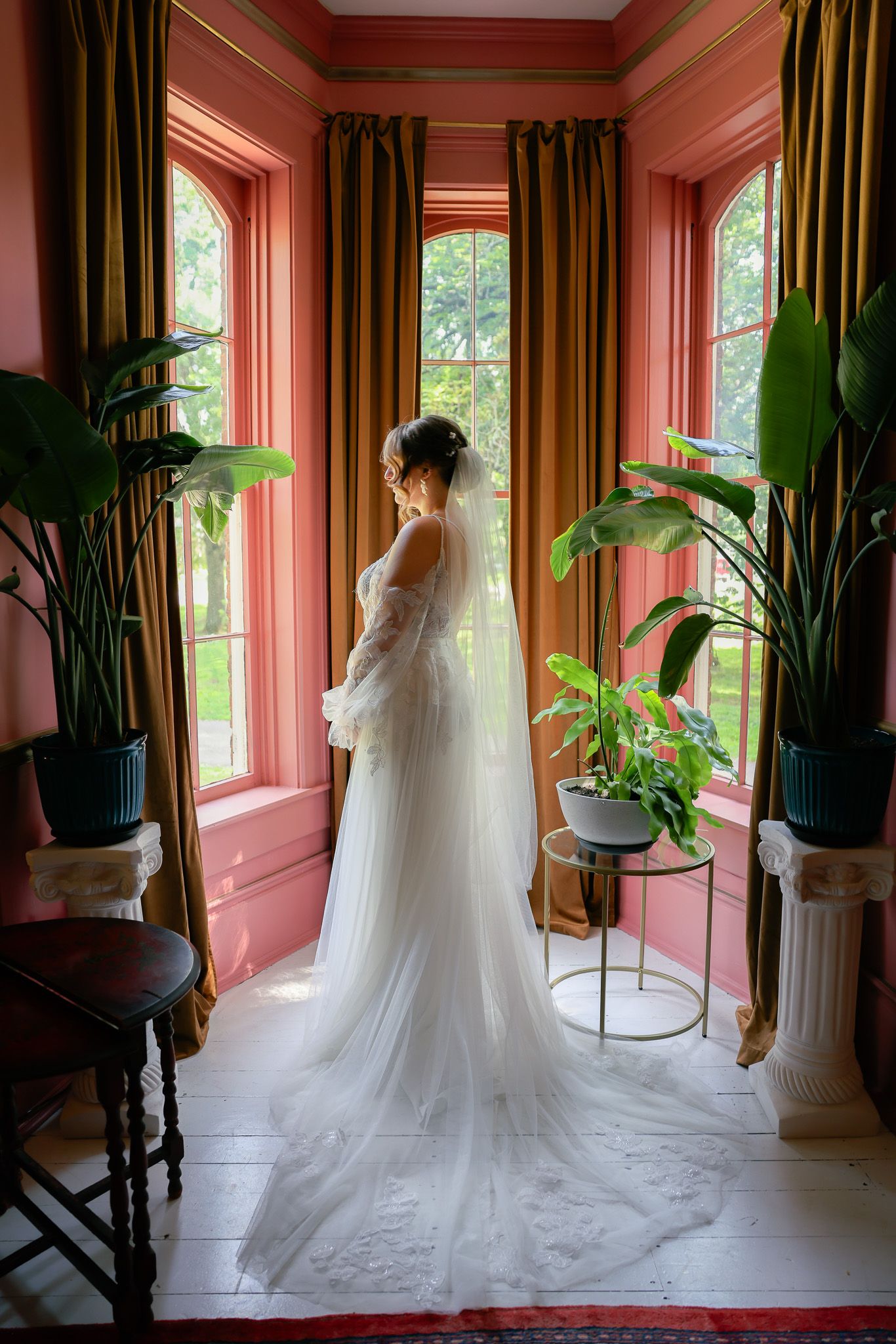 A flowing white wedding dress displayed in an elegant pink room with large windows and plants.