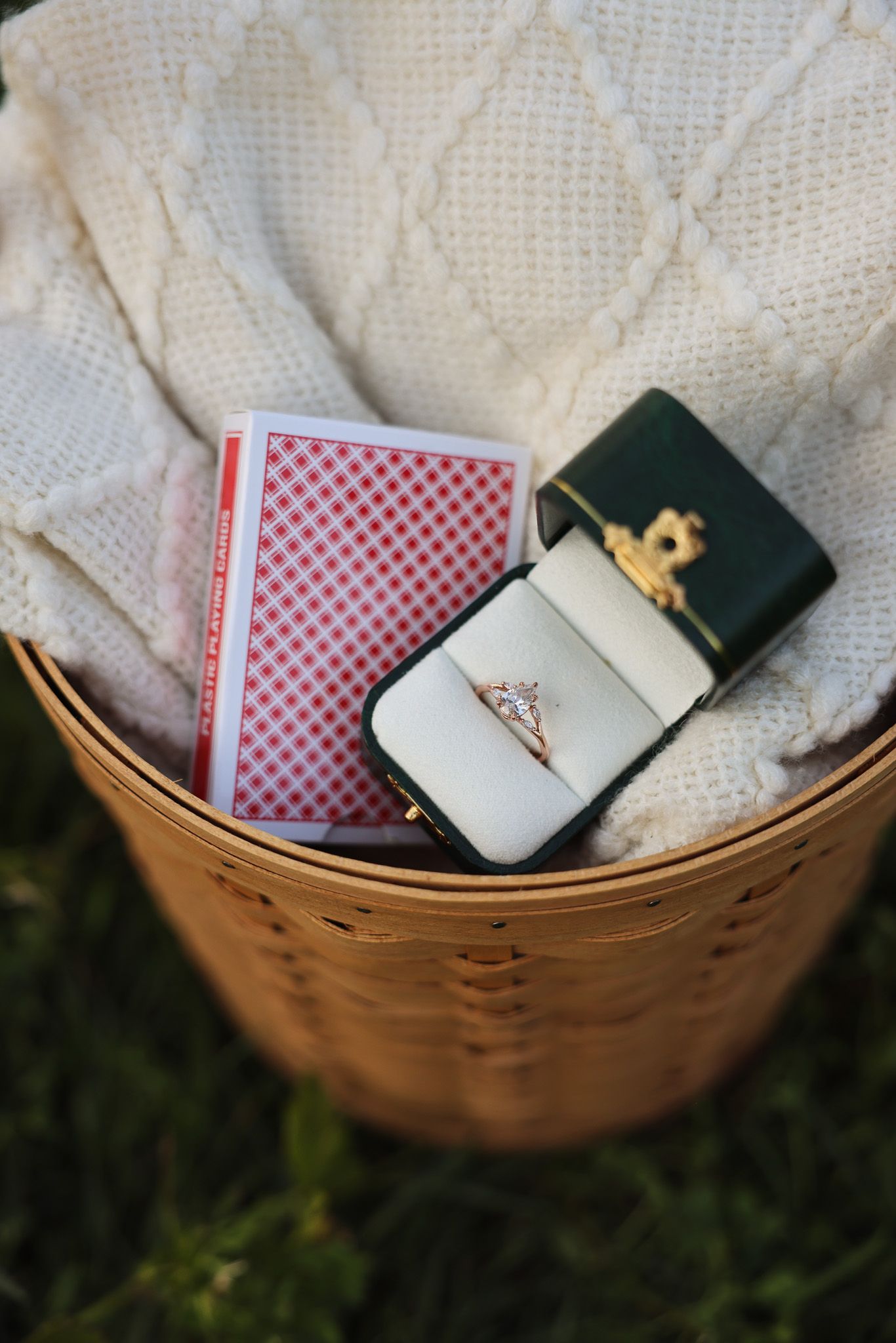 Woven basket containing ring box and red checkered playing cards on white fabric.