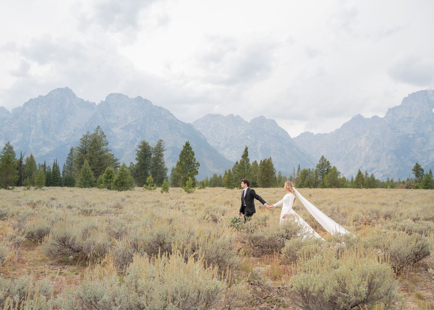 A wedding couple stands in a vast meadow with the Grand Teton mountains creating a dramatic backdrop.