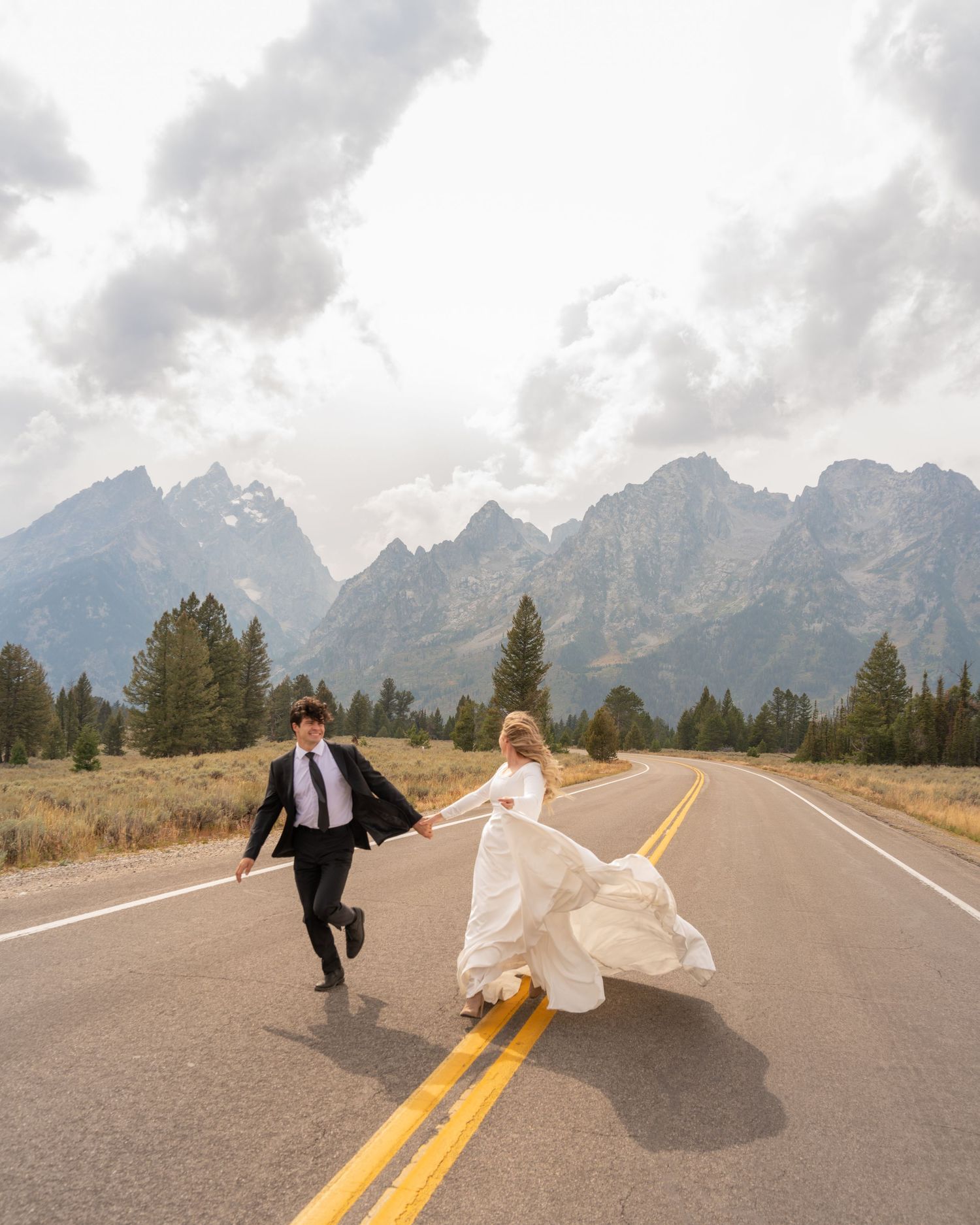 A couple in wedding attire playfully runs down an empty mountain road with the Grand Tetons in the background.