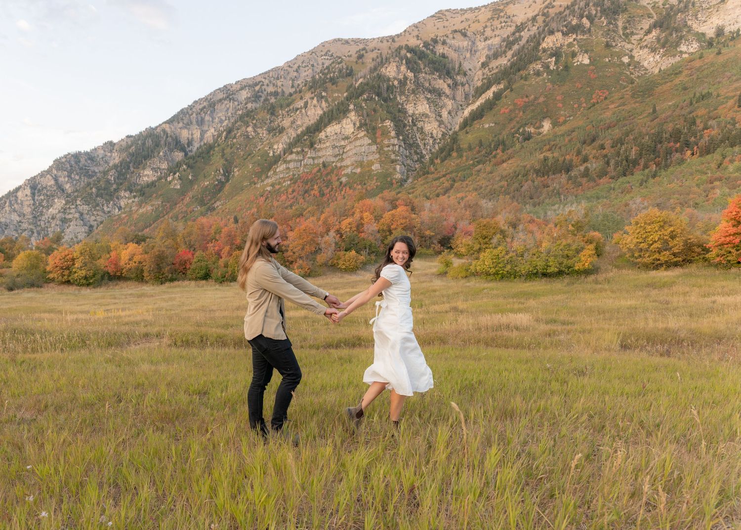 Couple walks hand in hand through a scenic mountain meadow during autumn with golden grass and fall foliage.