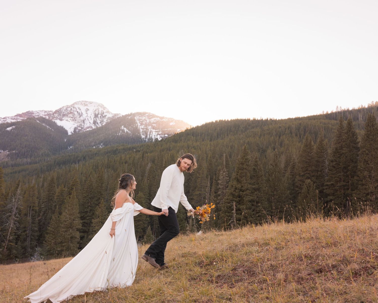 A couple in wedding attire walks through a mountainous landscape with snow-capped peaks and evergreen forests.