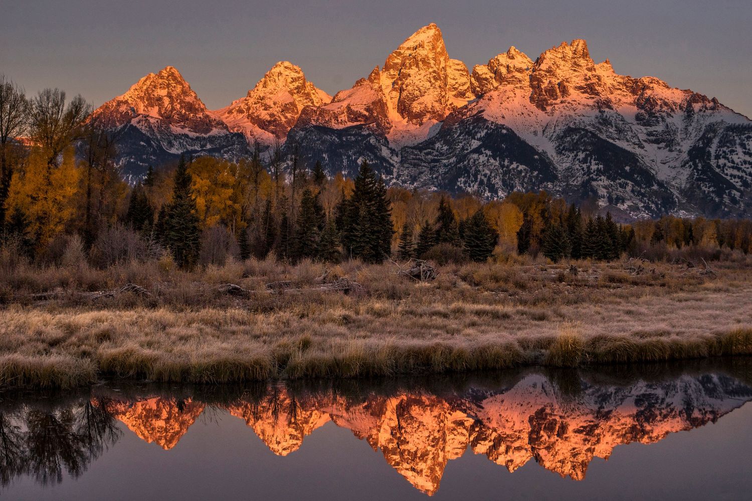 The Grand Tetons glow orange at sunrise, perfectly reflected in still waters with autumn trees.