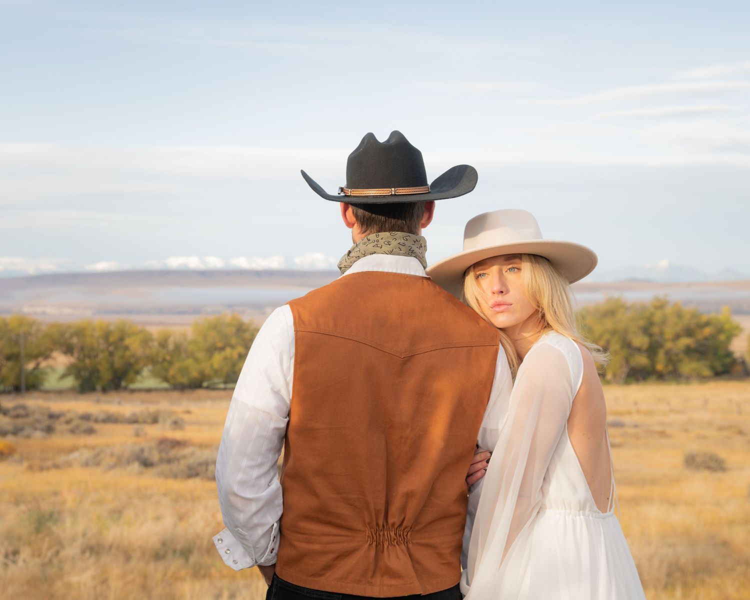 Western-styled couple in cowboy hats standing in prairie field at sunset.