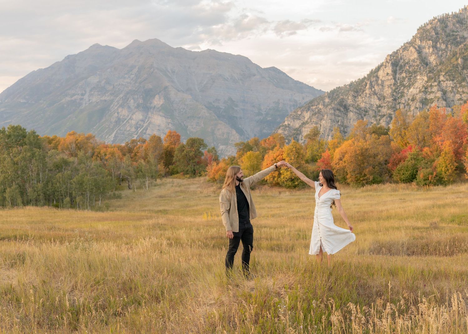 A couple stands together in a scenic mountain meadow during autumn with golden grass and dramatic peaks in the background.