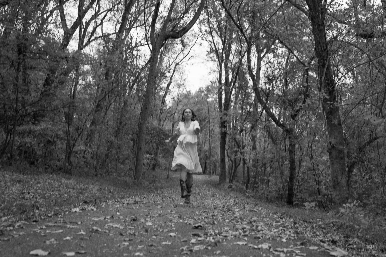 A figure in a white dress runs through a wooded forest path in black and white photography.