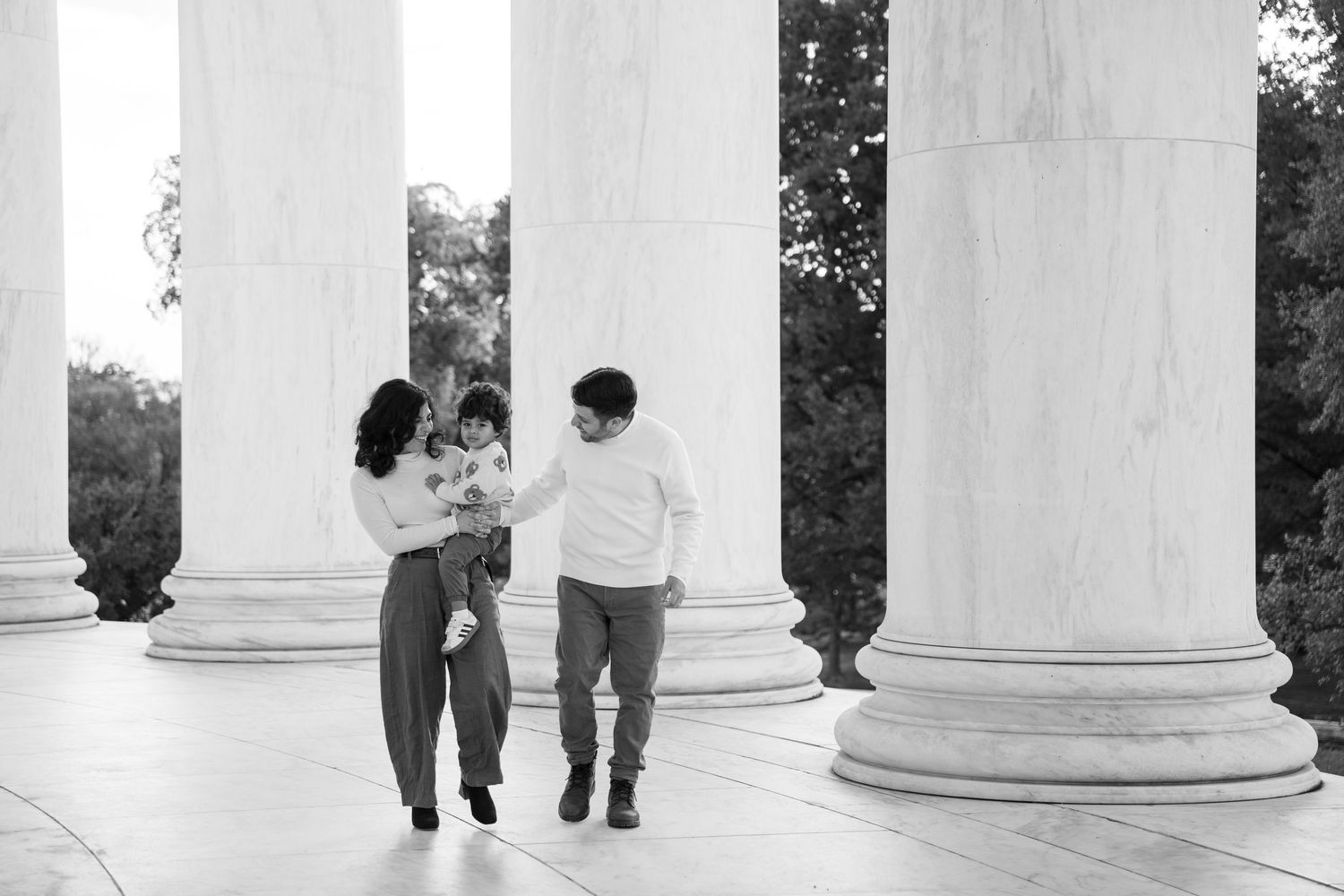 Black and white photo of a couple walking between classical white columns while holding a small child.