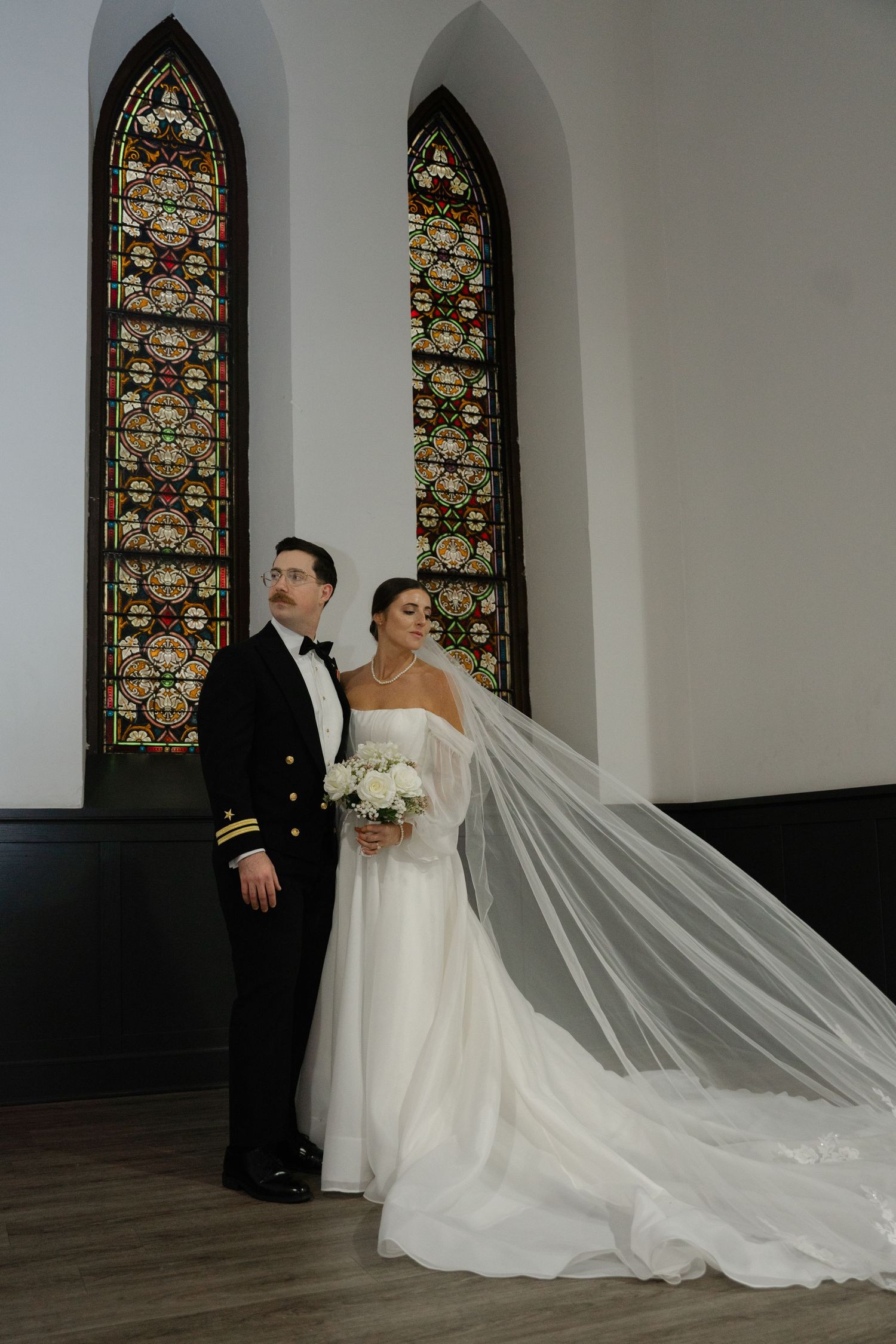 A couple poses in front of stained glass church windows during their wedding ceremony, with a flowing white veil trailing behind.