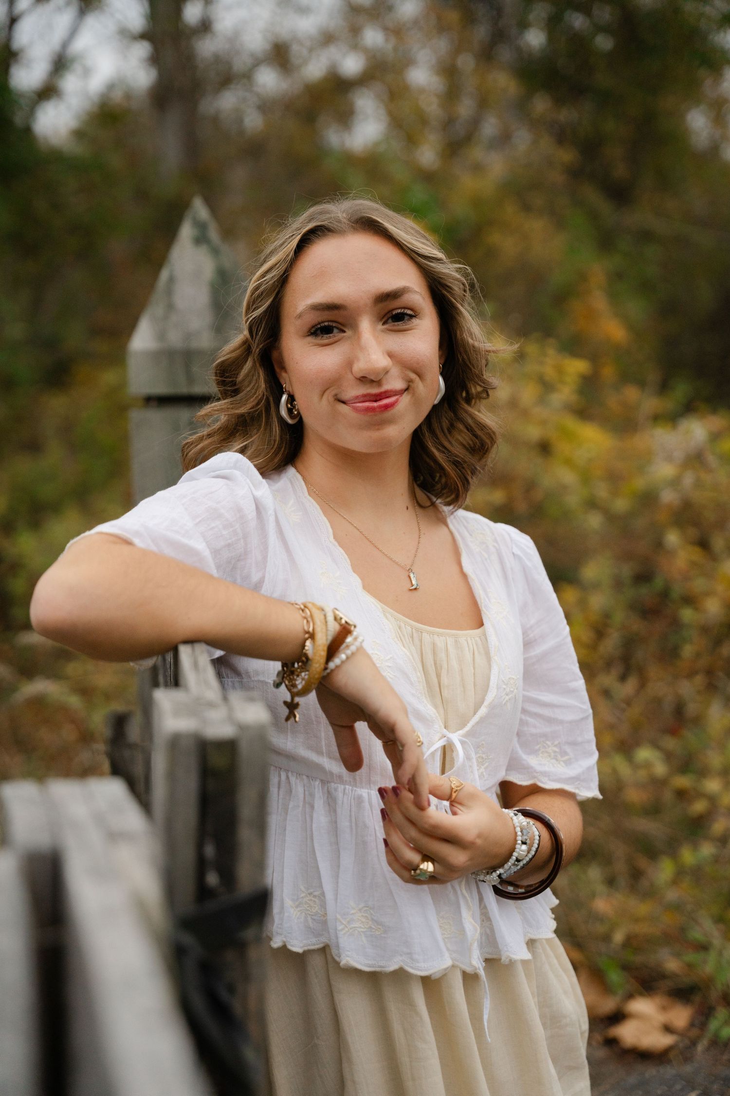 A person in a white peasant blouse poses outdoors in an autumn setting with jewelry and bracelets.