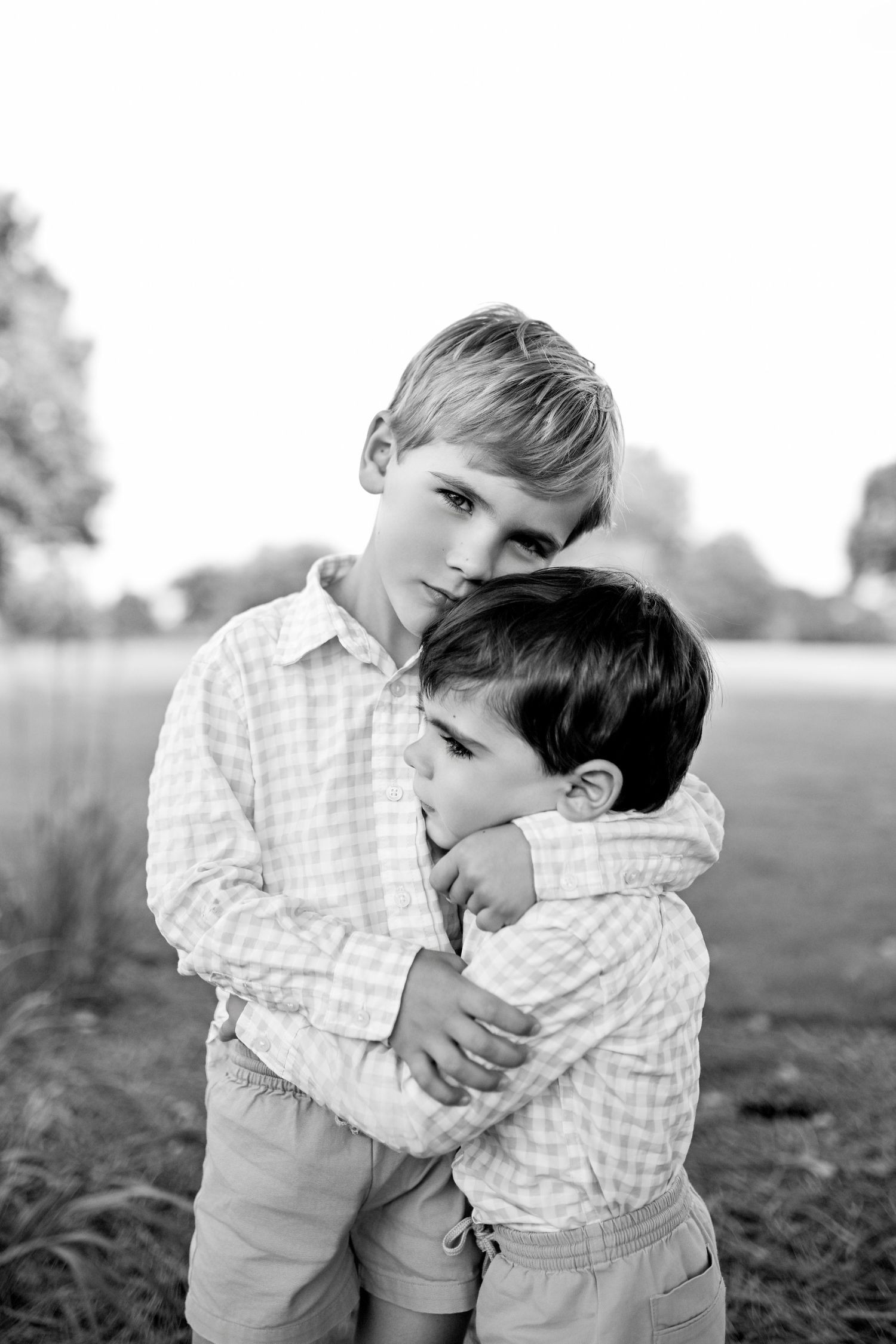 Two children in matching gingham shirts share a tender embrace in a series of black and white outdoor portraits.