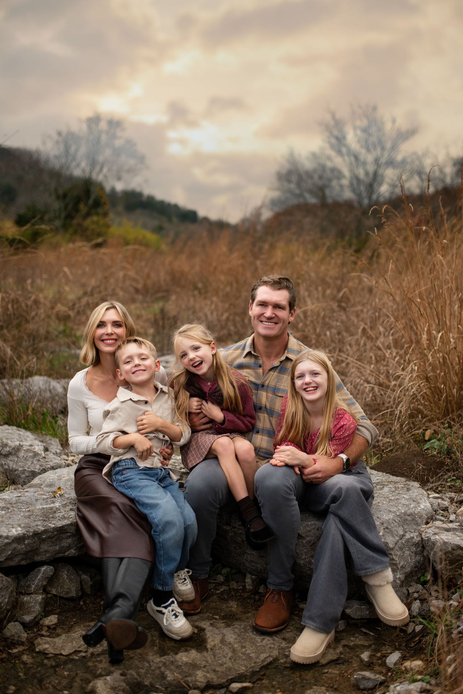 A family sits together on large rocks outdoors during autumn with a cloudy sky and brown foliage in the background.
