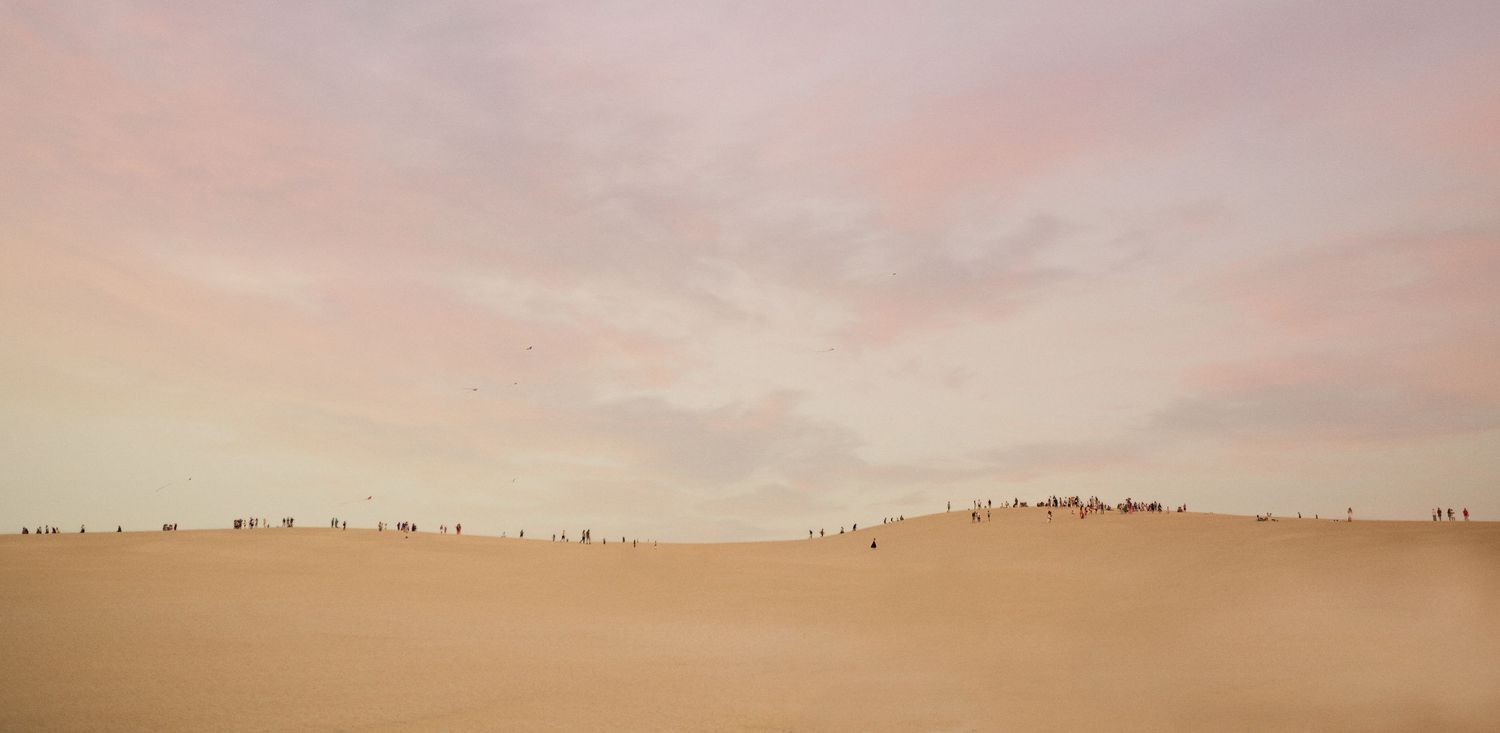 Vast sandy desert dunes under a soft pink and peach sunset sky with tiny silhouettes of people along the ridge.