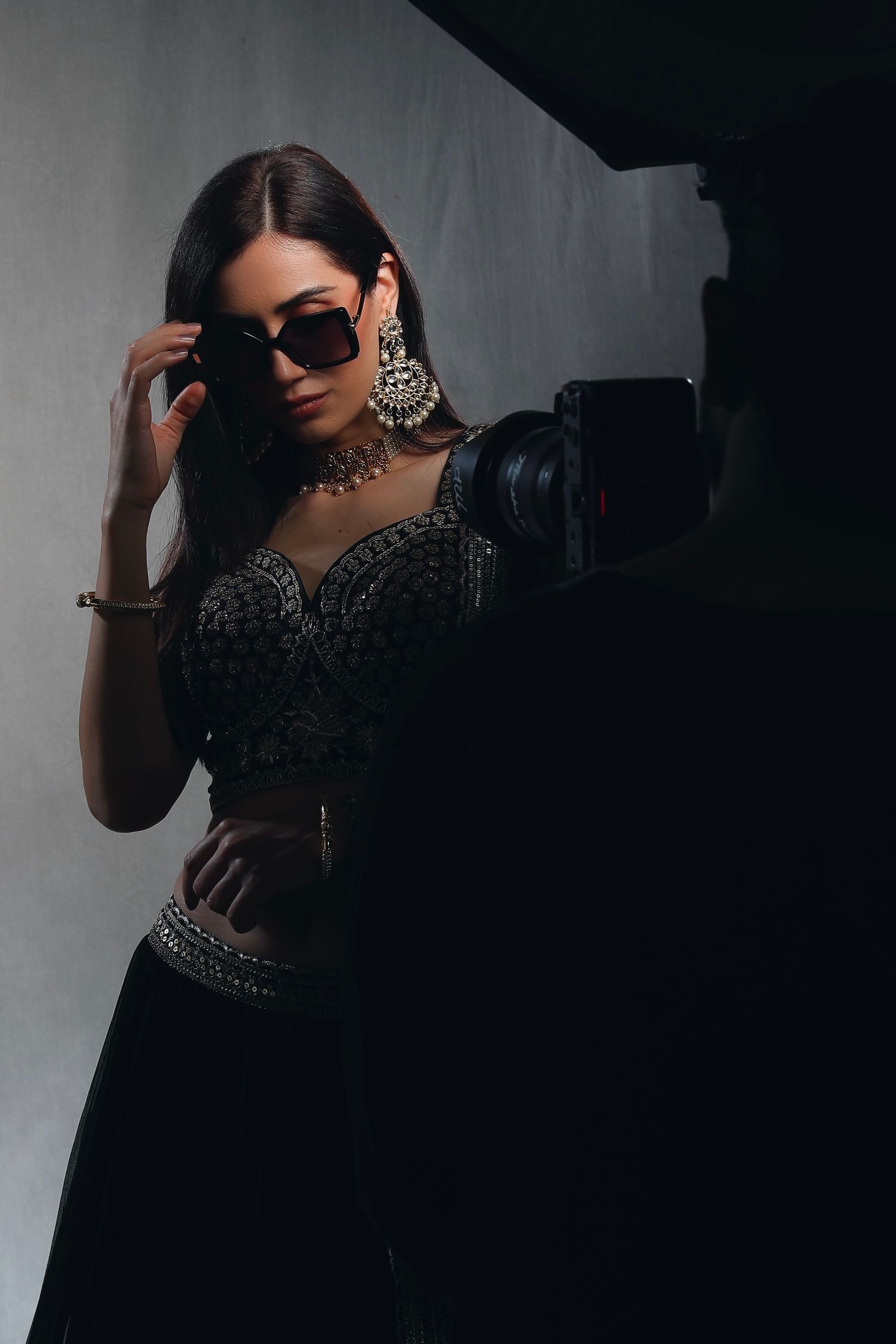Woman in black embroidered lehenga and silver jewelry adjusts sunglasses during a studio photoshoot.
