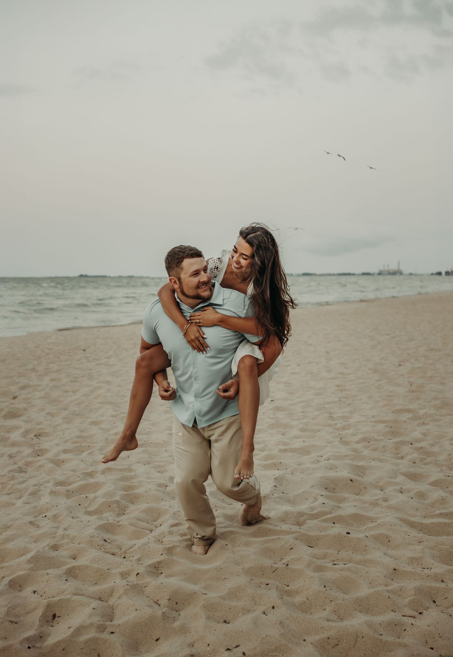 A couple shares a playful piggyback ride moment on a cloudy beach at sunset, creating a romantic coastal scene. Beach engagement