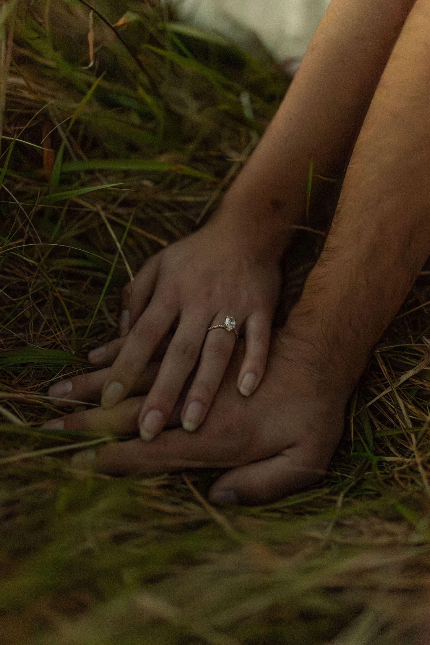 Close-up of hands with engagement ring resting on grass in moody natural lighting.